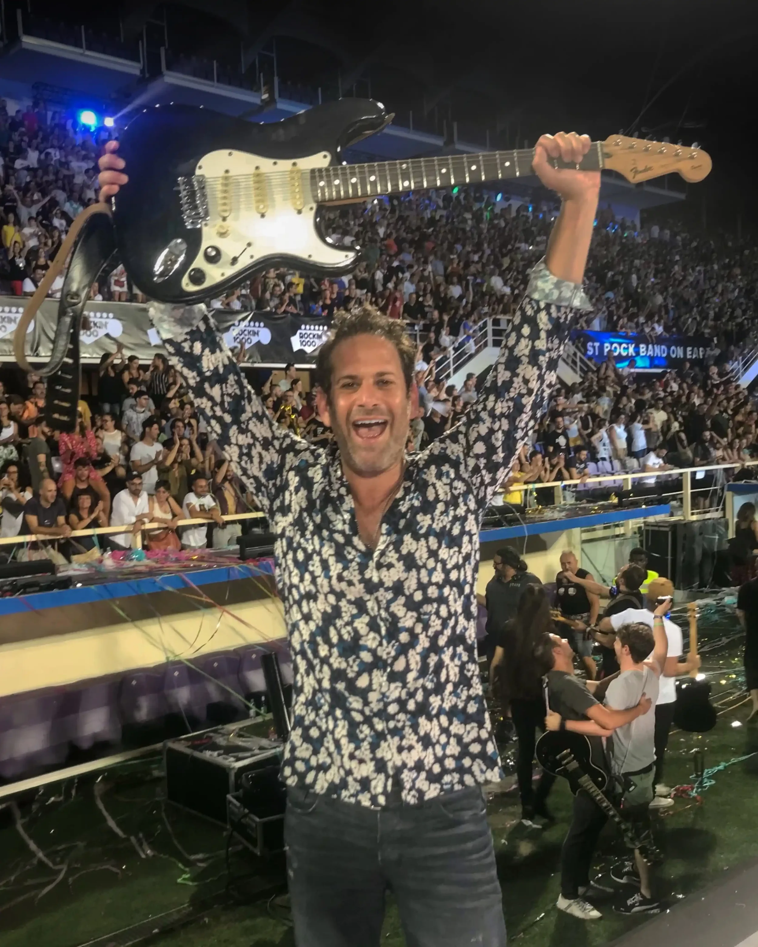 A man in a floral shirt holding a black electric guitar with a crowd in the background at an indoor concert or event.
