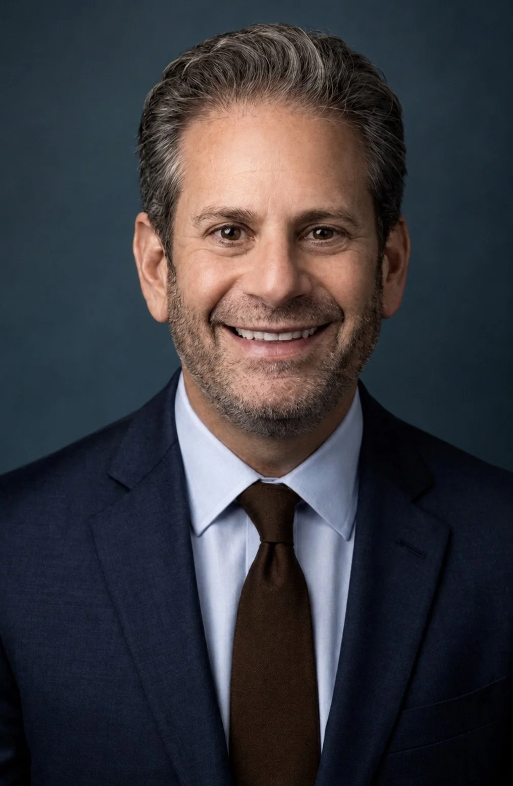 Professional headshot of a middle-aged man with gray hair and a beard, wearing a dark suit, white shirt, and brown tie, smiling against a dark background.