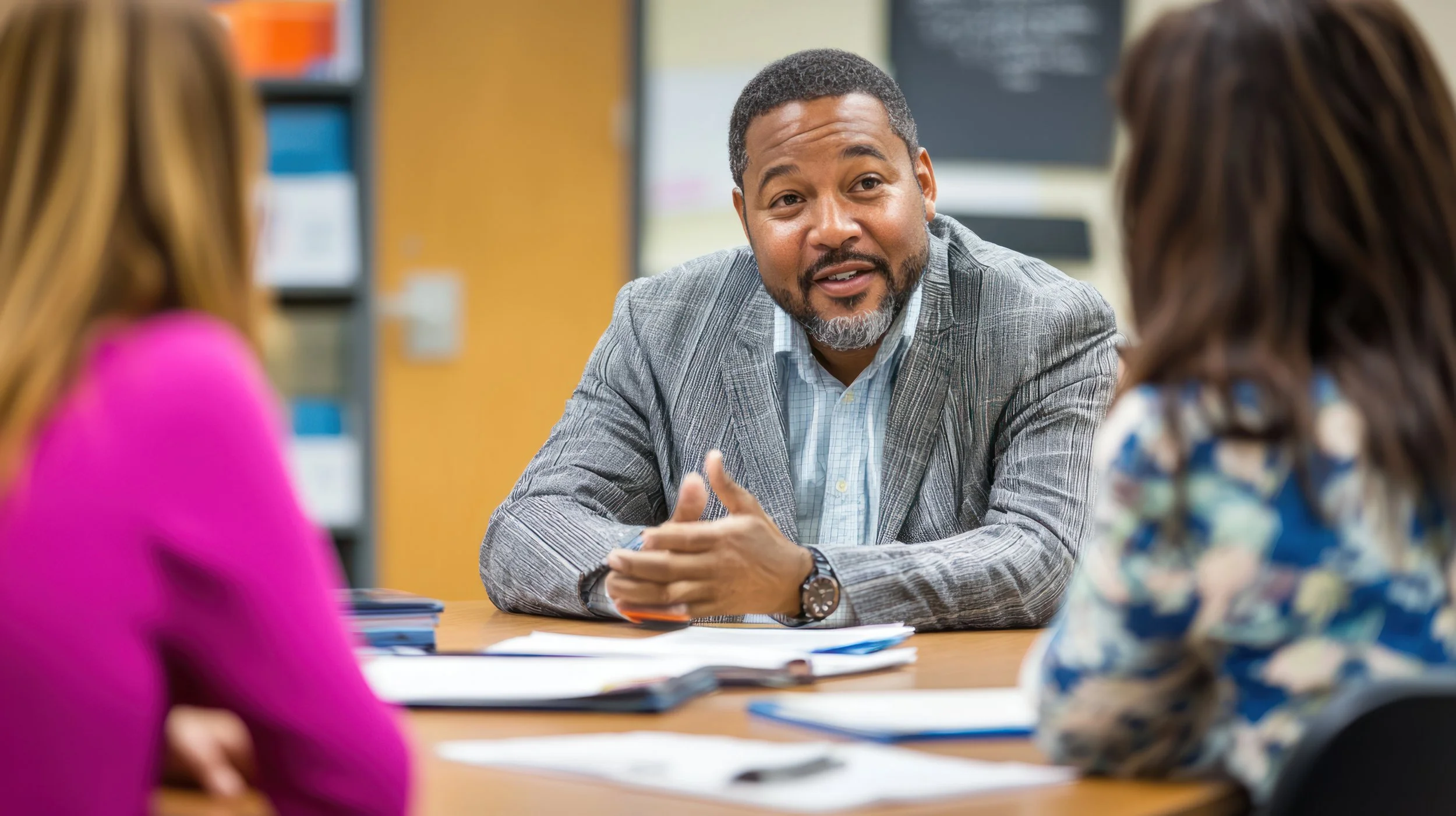 A man with a beard and short hair, wearing a gray suit jacket and light blue shirt, speaking to two women in a classroom or office setting with books and paperwork on the table.