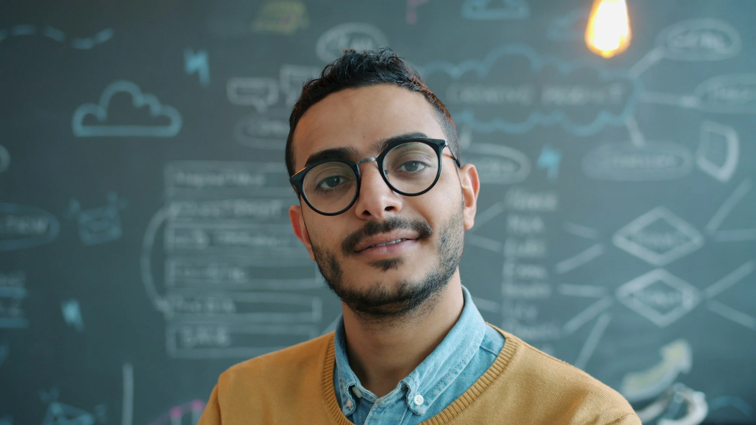 A young man wearing glasses and a mustard sweater standing in front of a blackboard filled with chalk drawings of clouds, arrows, and text.