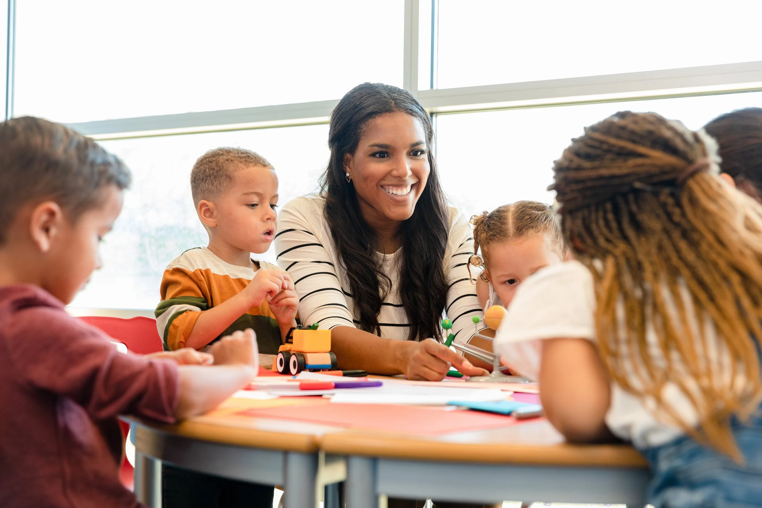 A woman with long dark hair and earrings, smiling, sitting at a table with young children engaged in a classroom activity. The children have varied hair styles and are focused on their work, with some toys and supplies on the table. Large windows in the background let in natural light.