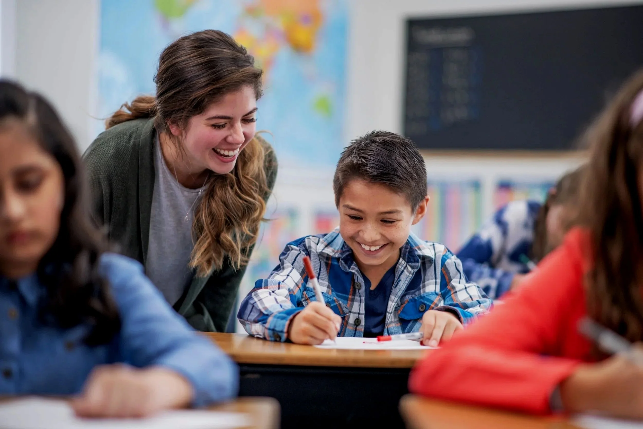 A smiling teacher helps a young boy with a writing assignment in a classroom. Other students are blurred in the foreground.