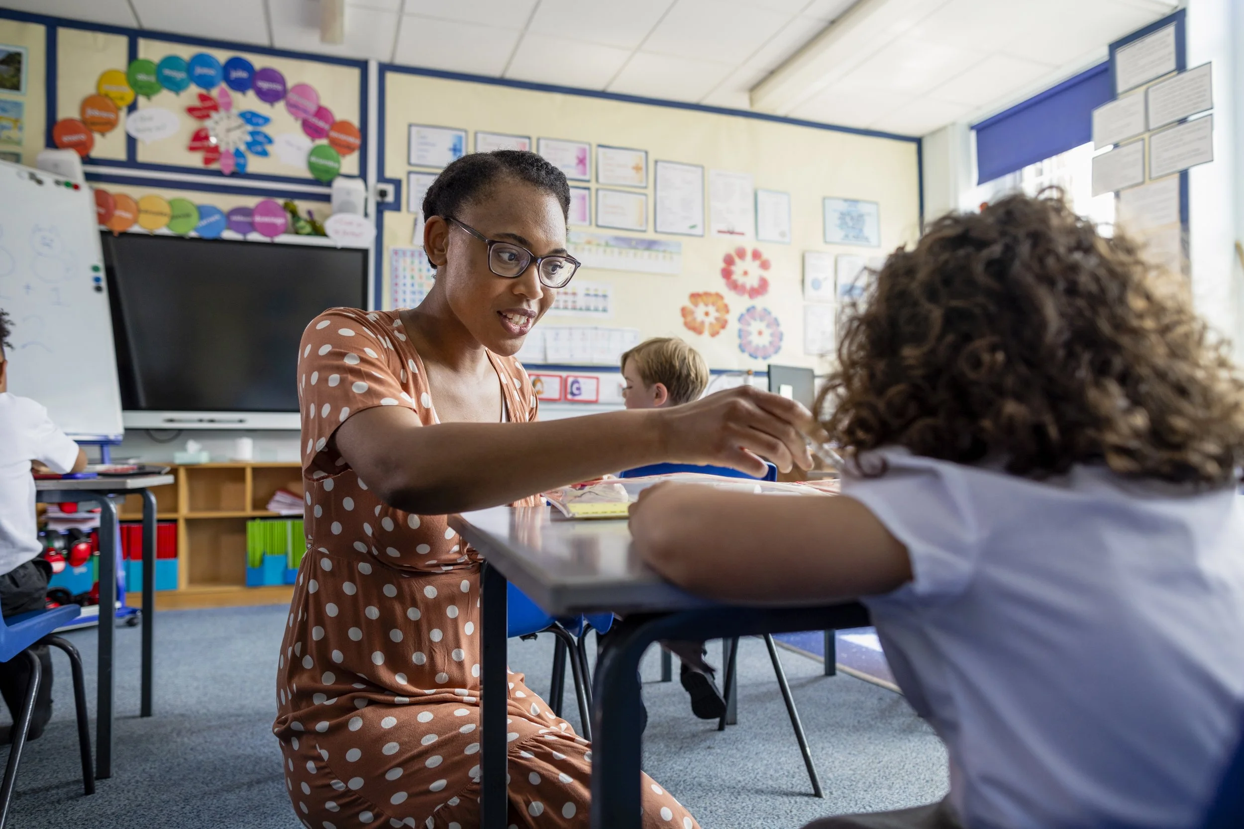 A classroom scene with a teacher in a polka dot dress engaging with a student at the desk, other children present, decorated educational posters, and a whiteboard.