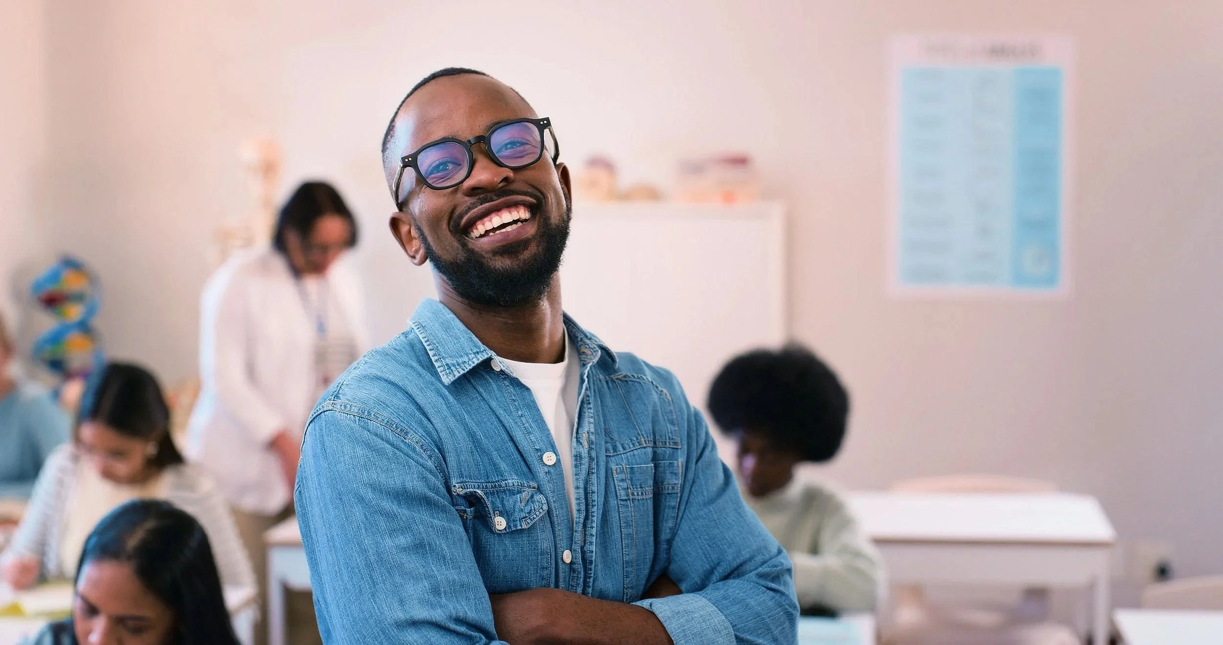 A smiling man with glasses and a denim shirt stands with arms crossed in a classroom filled with students, some working at desks and a teacher in the background.