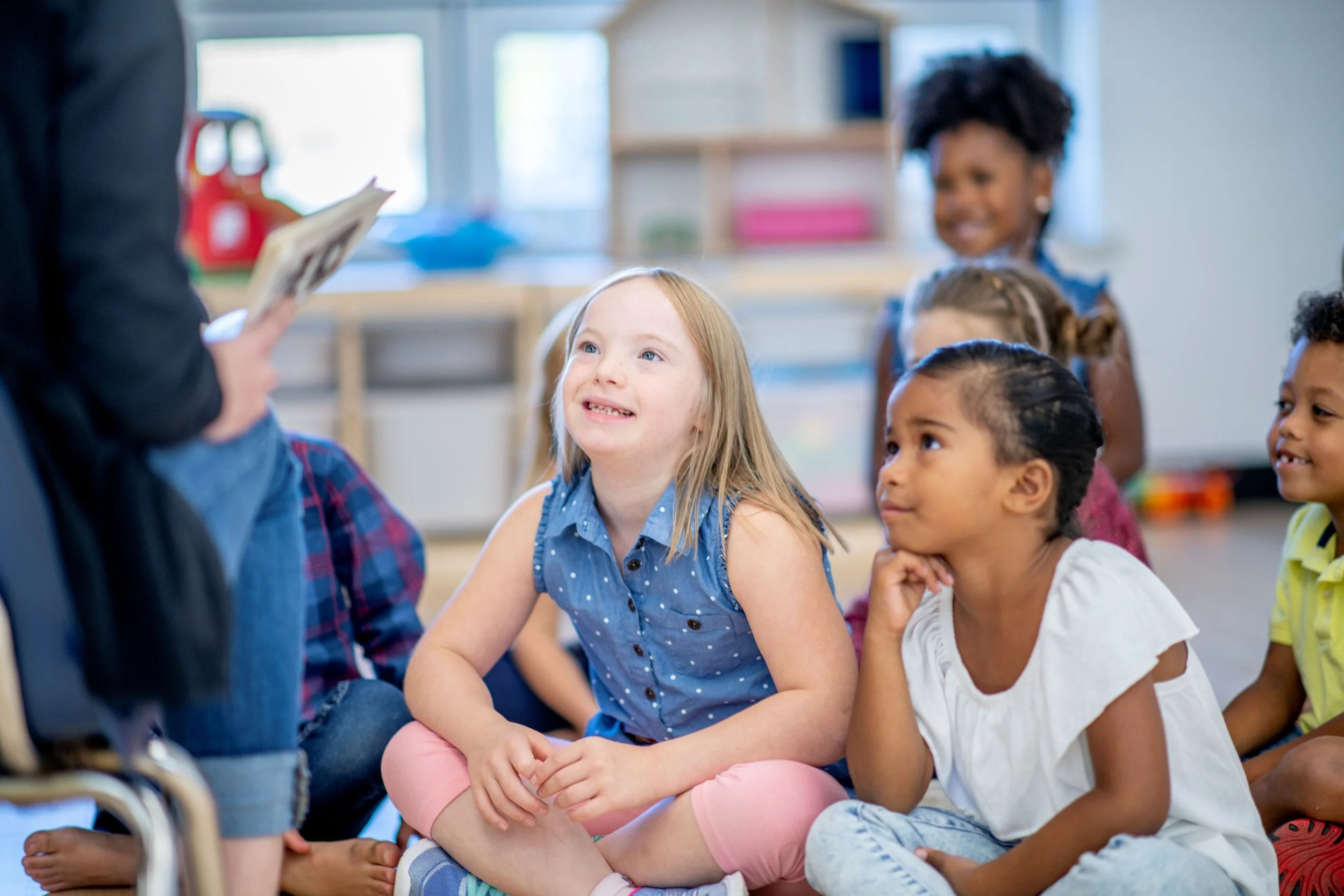 A group of young children sitting on the floor and listening attentively to an adult reading a book, inside a brightly lit classroom.
