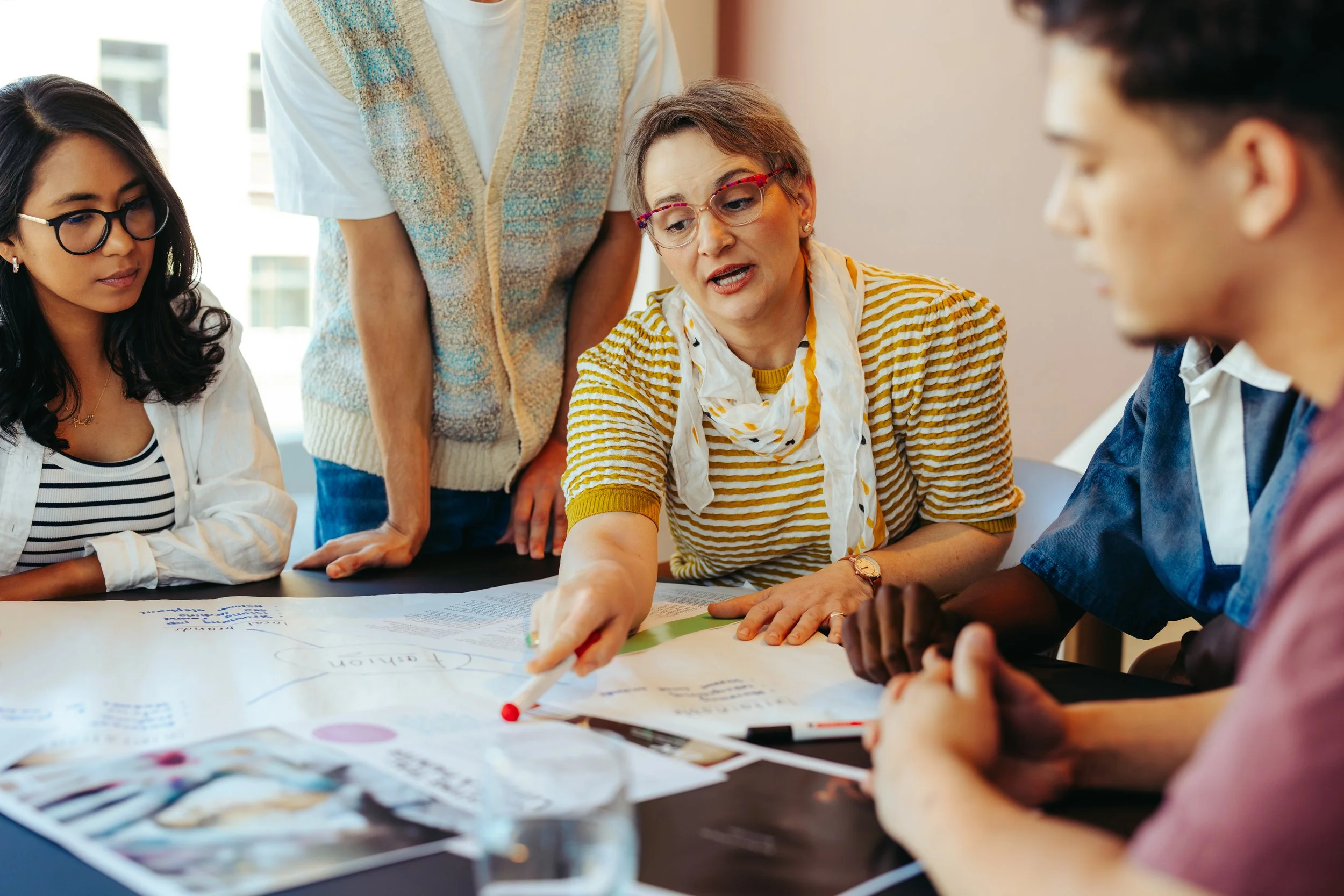A diverse group of people in a meeting, focusing on a woman with glasses and short hair explaining something on a large paper on the table while others listen and look on.
