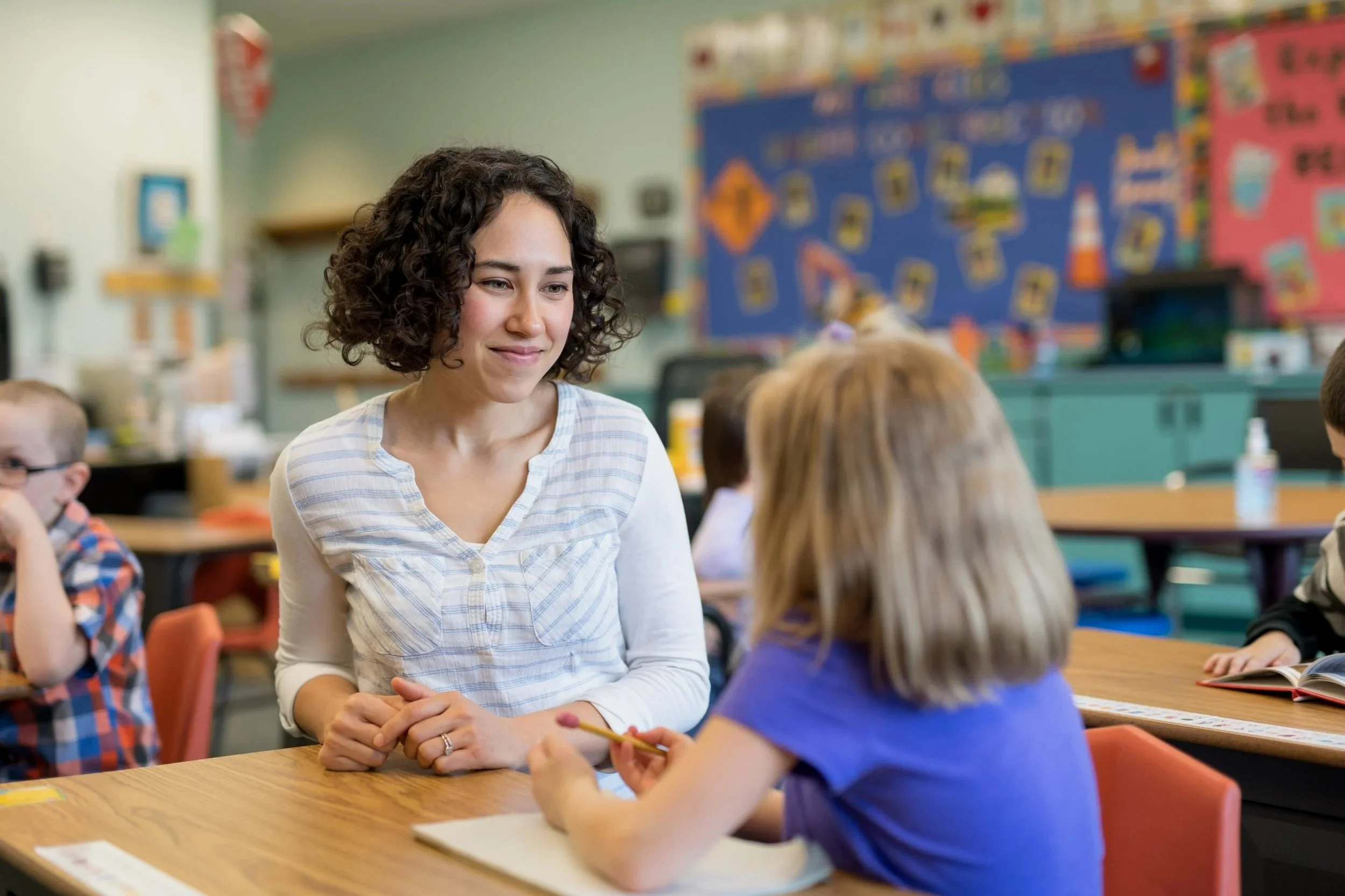 A teacher with curly brown hair in a white checkered shirt smiling and talking with a young student wearing a blue shirt in a colorful classroom with children and educational decorations.