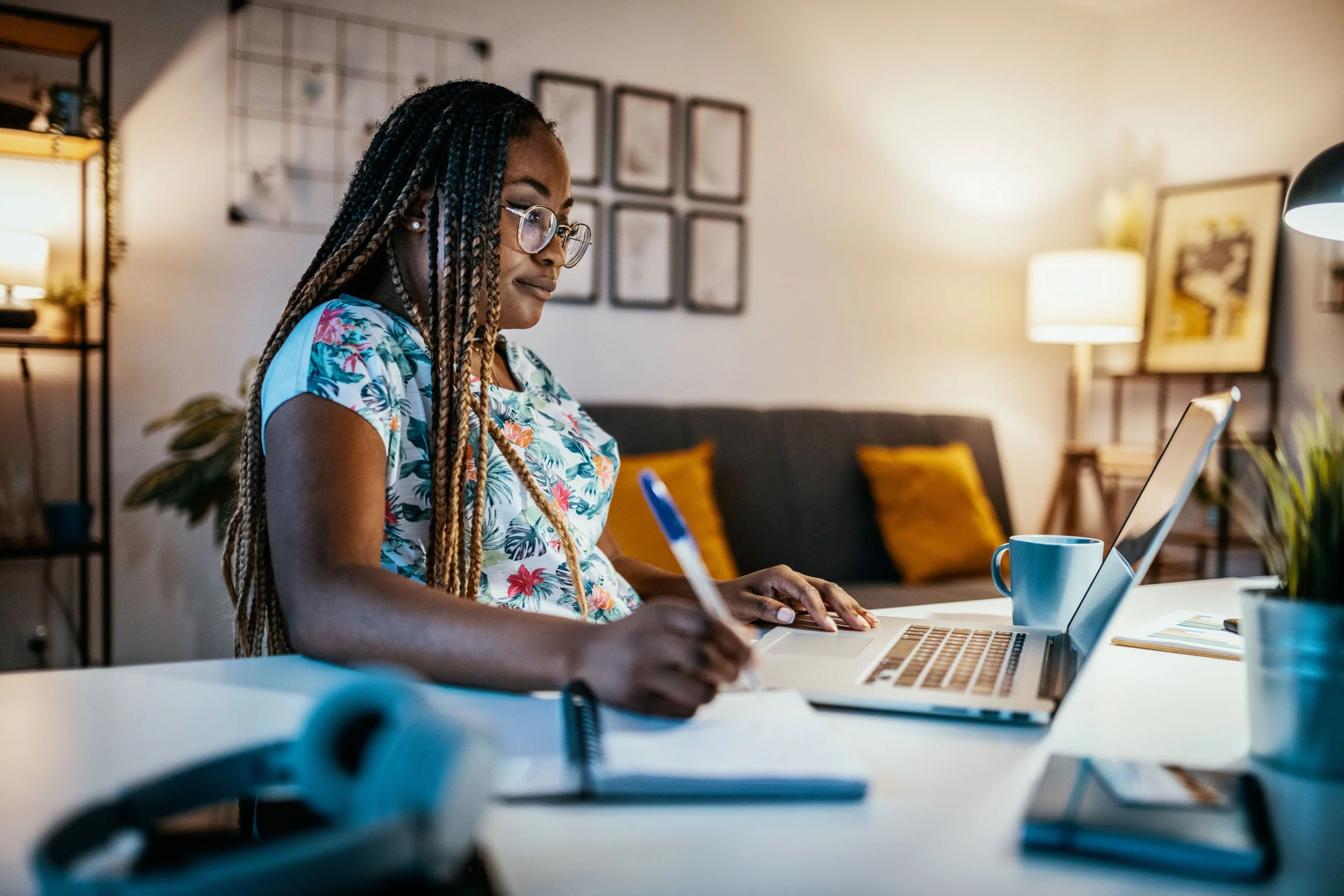 A woman with long braided hair, wearing glasses and a patterned shirt, working on a laptop at a desk in a cozy living room. The background features a black sofa with yellow cushions, picture frames on the wall, and a standing lamp.