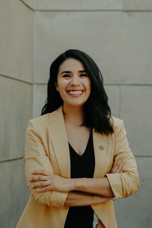 A woman with shoulder-length black hair smiling and crossing her arms, wearing a beige blazer over a black top, standing against a grey stone wall.