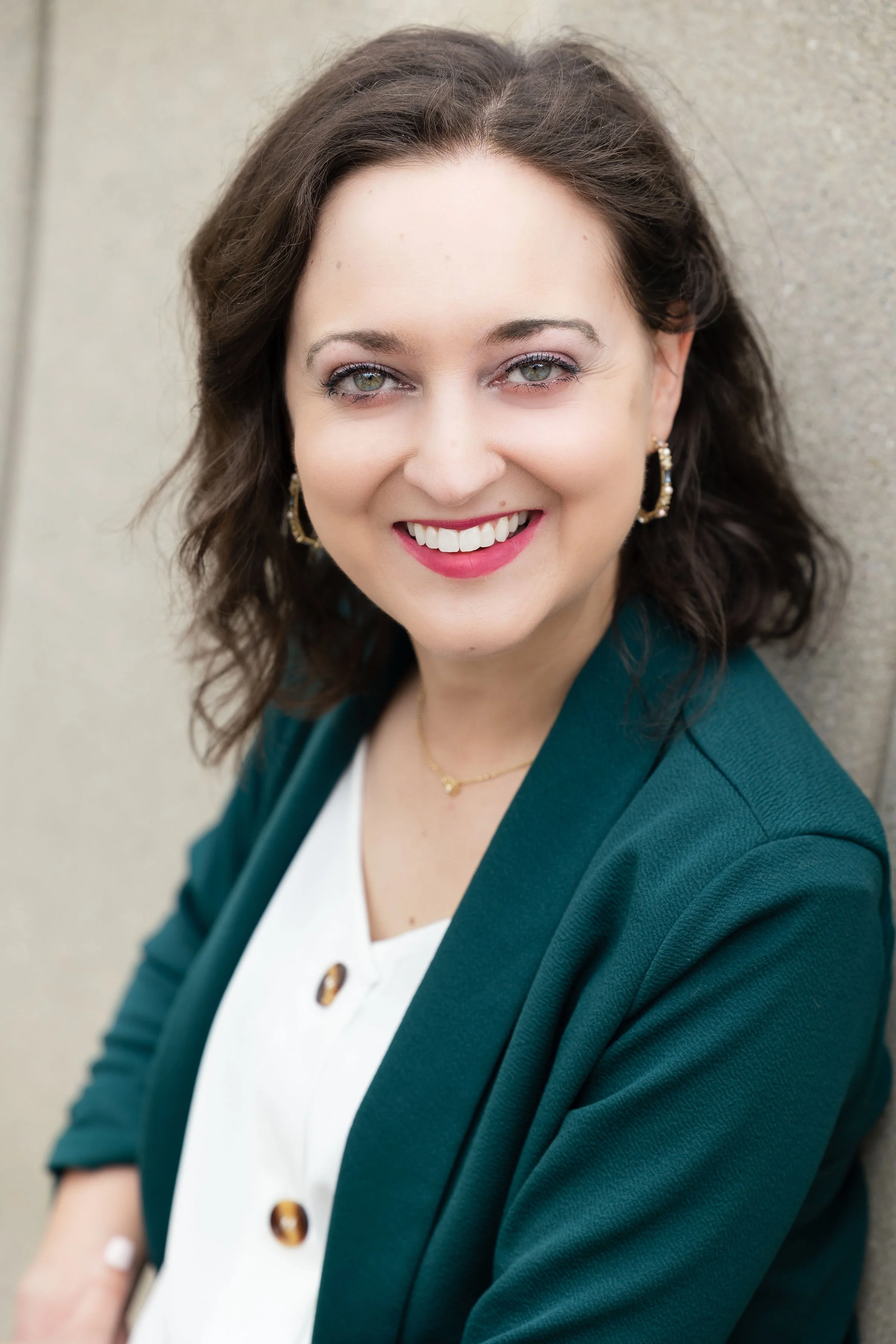 A woman with medium-length curly brown hair, wearing a dark teal blazer, a white blouse with large decorative buttons, gold hoop earrings, a delicate gold necklace, and pink lipstick, smiling and standing against a beige wall.