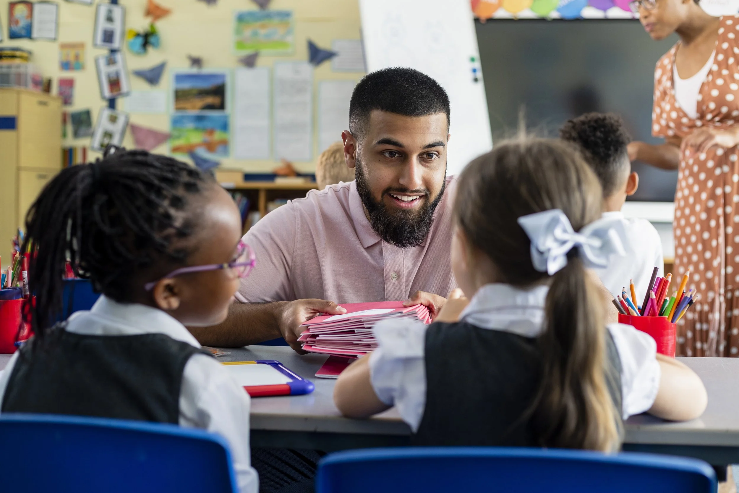 A teacher interacting with a group of young students in a classroom, with educational materials on the wall and a whiteboard in the background.