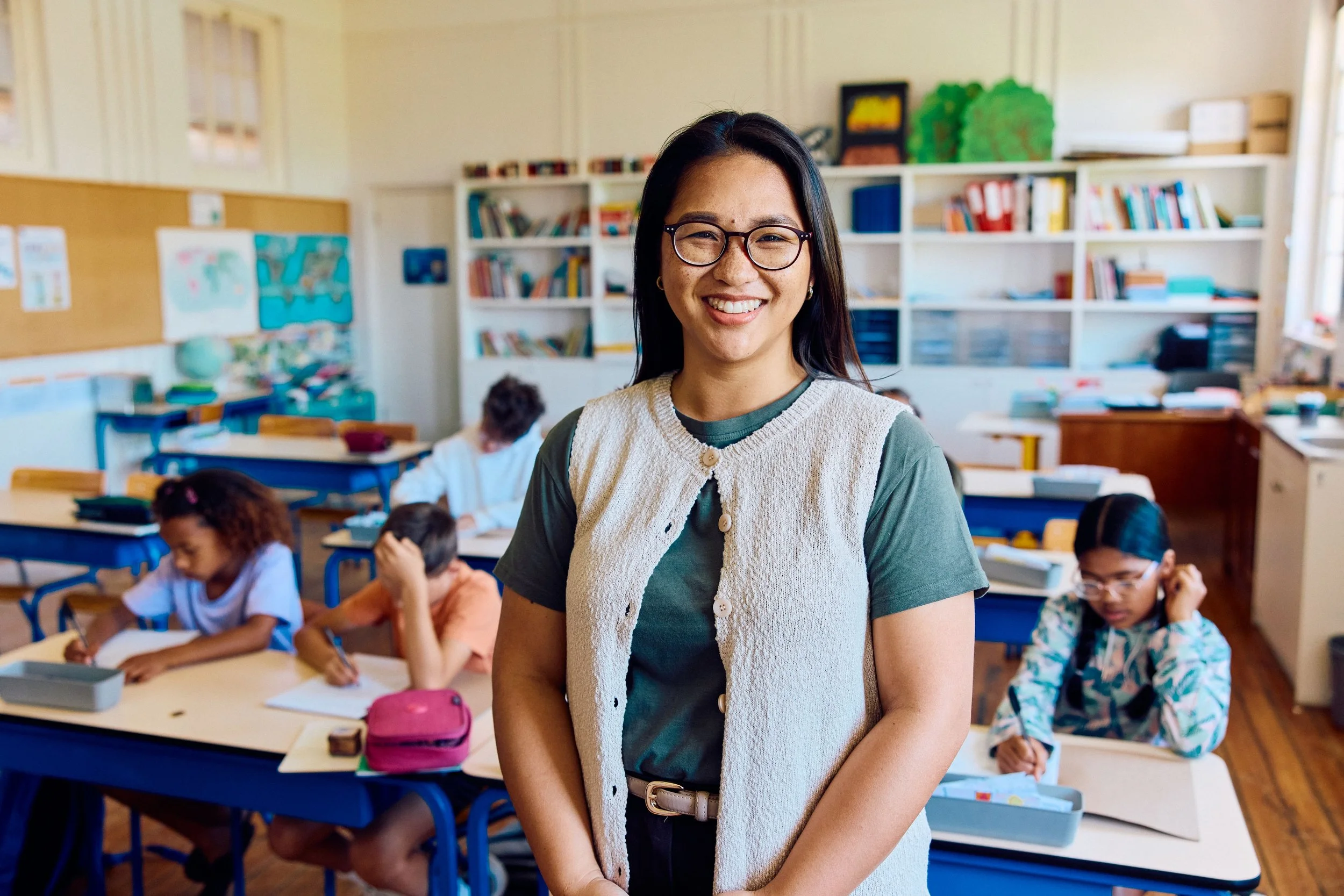 A smiling woman with glasses stands in a classroom filled with students working at desks. Bookshelves and educational posters are visible in the background.