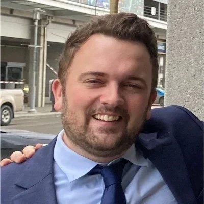 Portrait of a man with short brown hair and a beard, wearing a blue suit and tie, smiling outdoors in an urban setting.