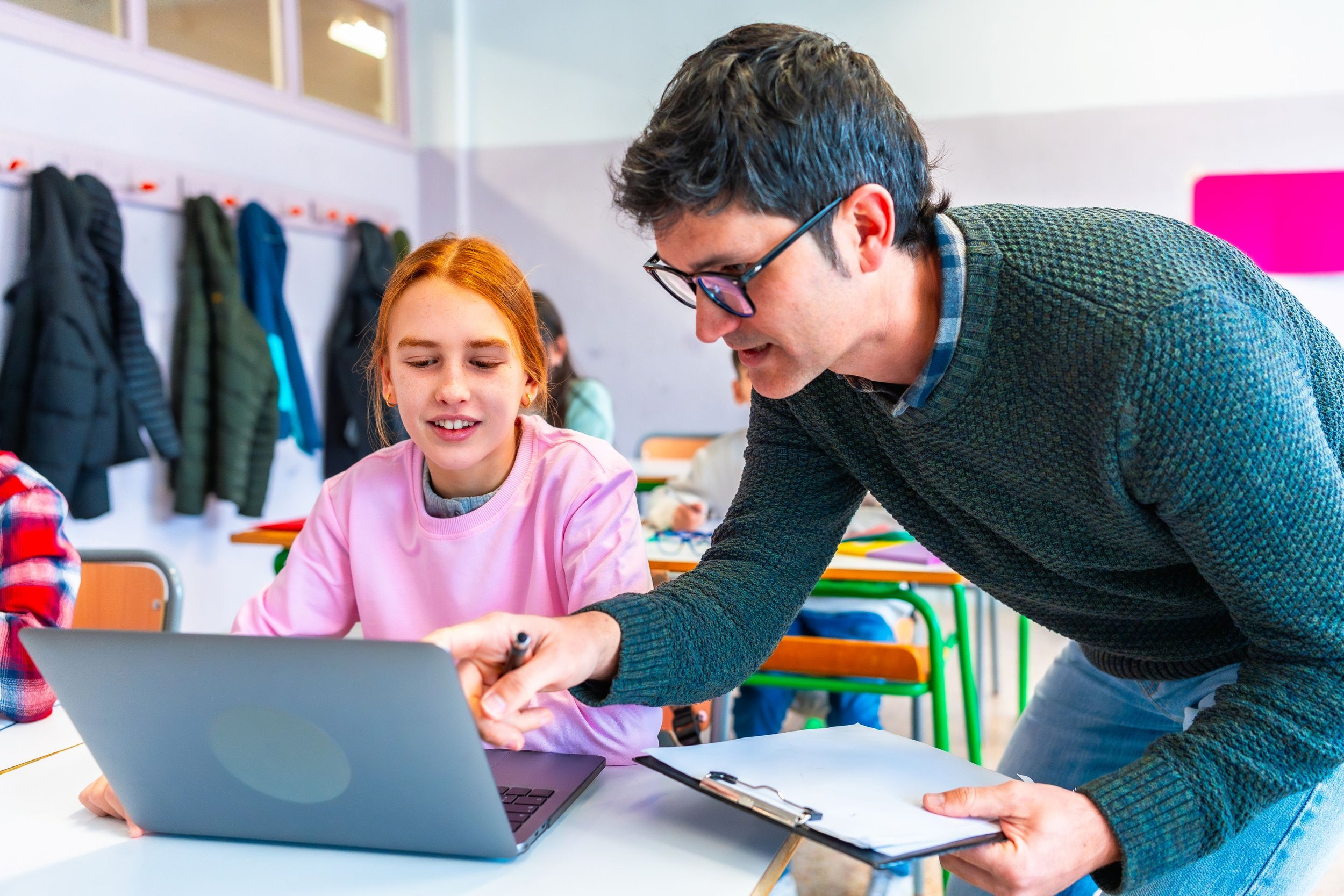A young girl with red hair wearing a pink sweater is seated at a desk in a classroom, looking at a laptop. An adult man, wearing glasses and a green sweater, is leaning over the desk, pointing at the laptop screen and holding a clipboard. The classroom has colorful chairs, hooks with coats on the wall, and other children in the background.
