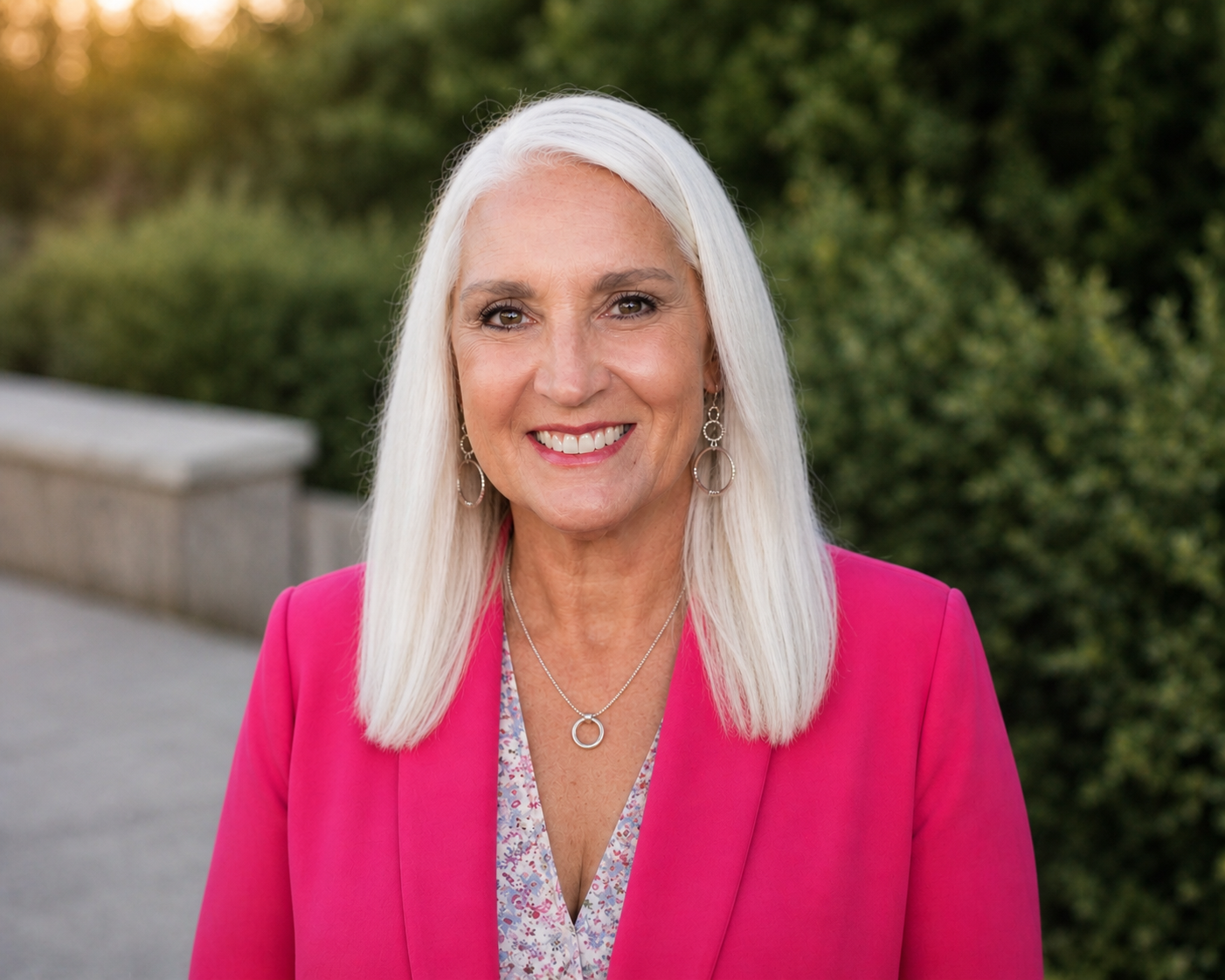 A smiling older woman with long, straight gray hair, wearing a bright pink blazer and jewelry, standing outdoors with greenery in the background.
