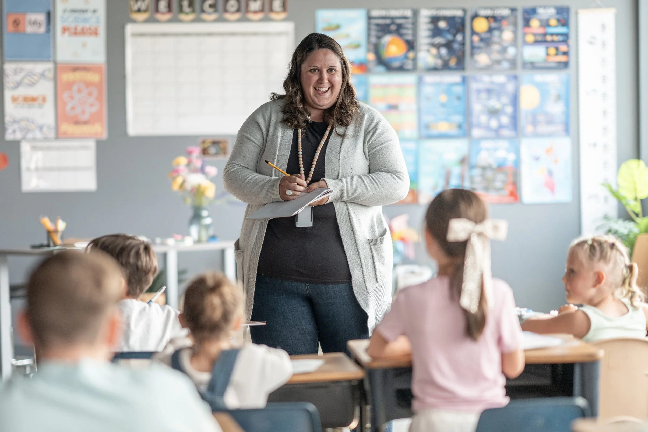 A smiling female teacher in a classroom with children, standing near a whiteboard and colorful posters on the wall.