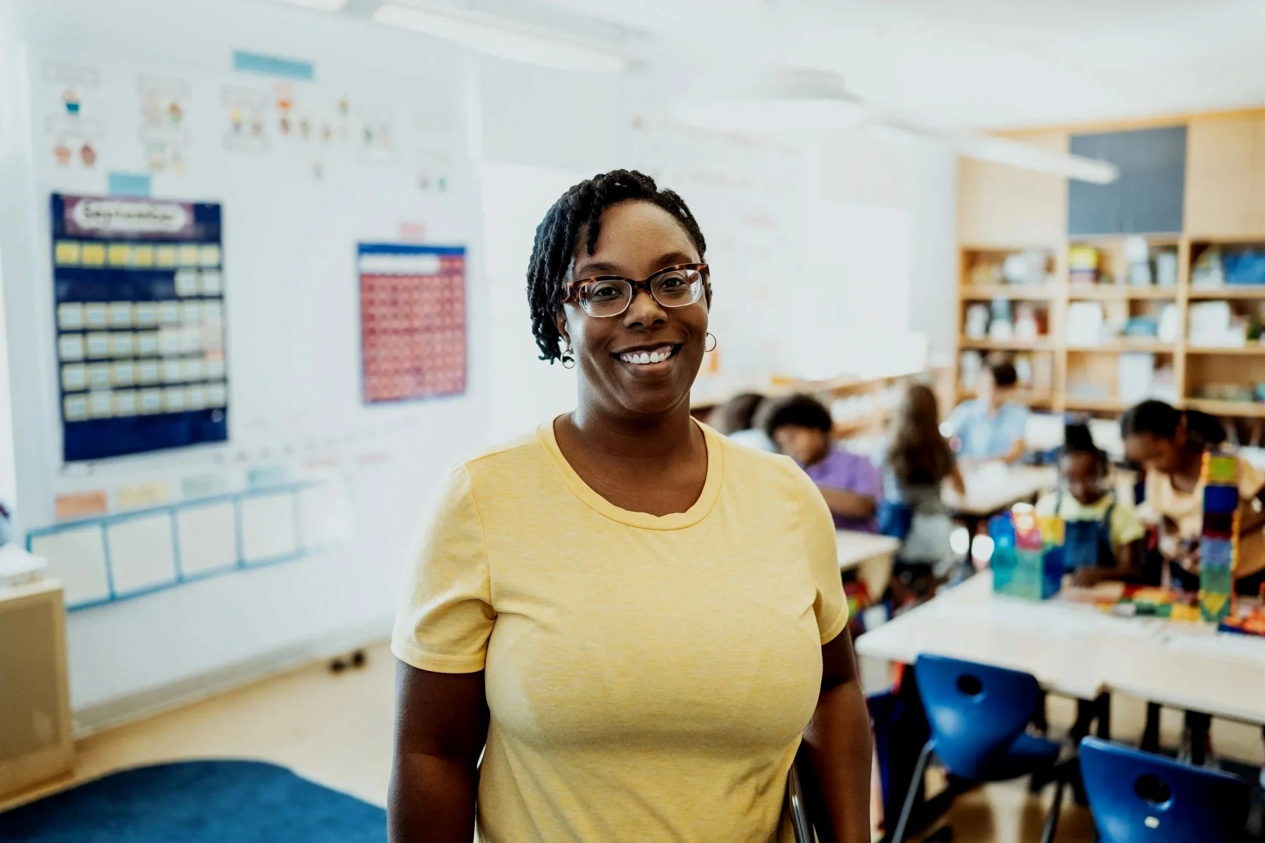 A smiling woman with glasses and short curly hair in a yellow t-shirt standing in a classroom with students in the background.