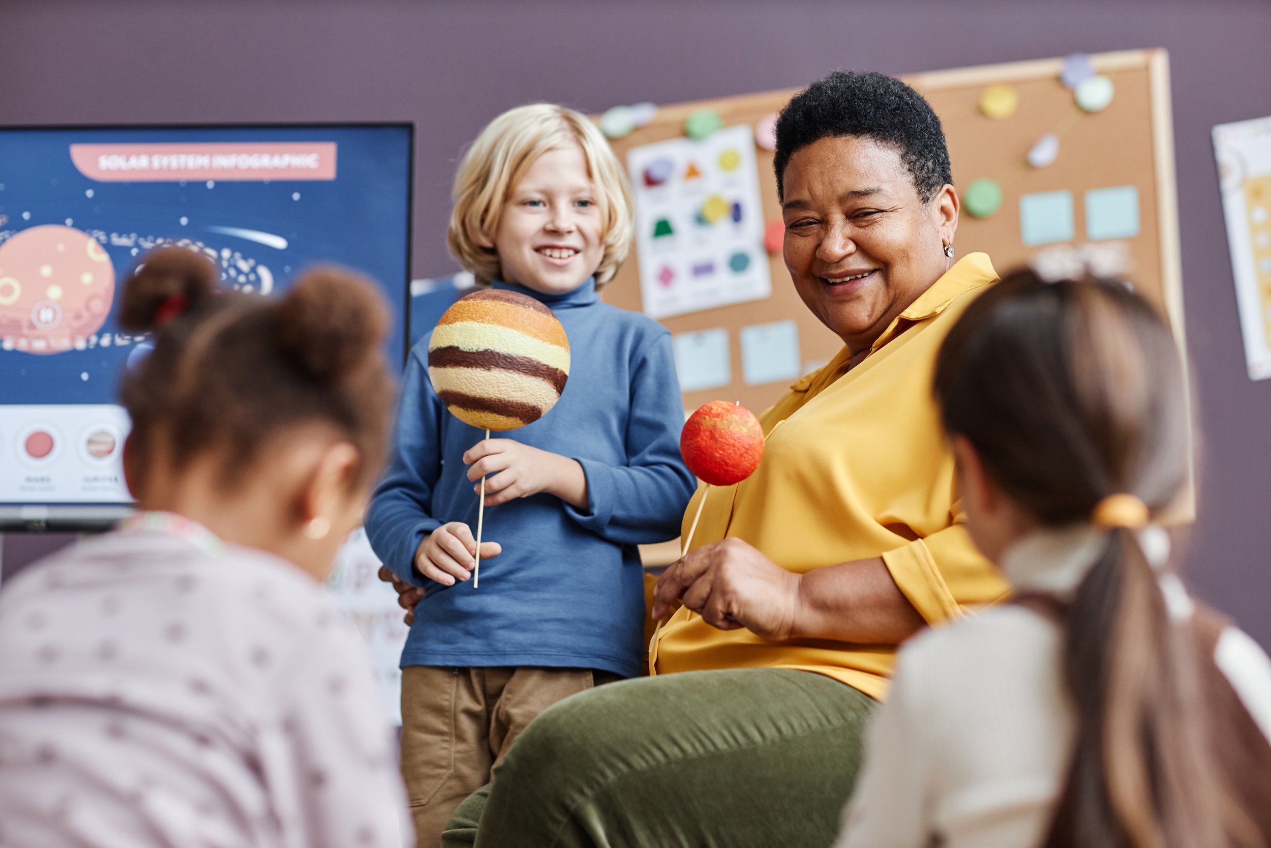 A teacher and a student holding models of planets during a classroom presentation, with other students seated and a solar system infographic on the screen in the background.