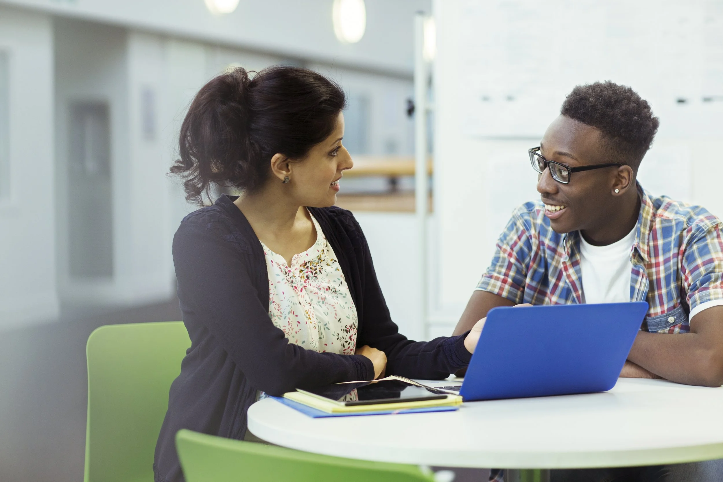 A woman and a young man sitting at a table, engaging in conversation. The woman has dark hair tied back and is wearing a floral blouse and dark blazer. The young man has short, curly hair, glasses, and is wearing a plaid shirt. They are in a bright, modern room with white walls and green chairs, with a laptop and notebooks in front of them.