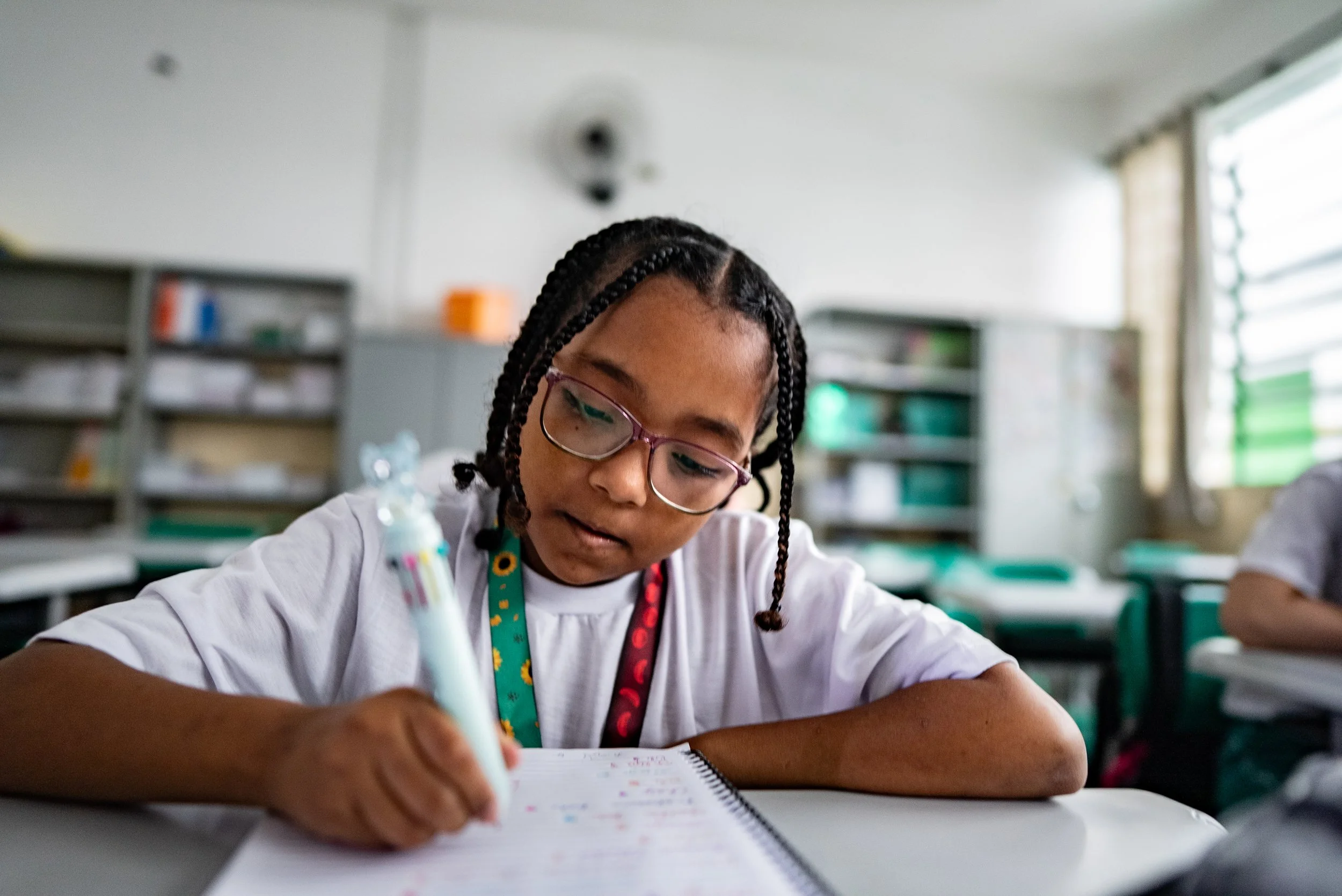 A young girl with braided hair and glasses writing in a notebook at a desk in a classroom.