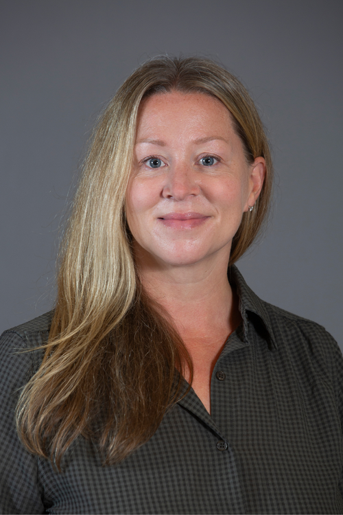 A woman with long blonde hair, wearing a dark checkered shirt, smiling in front of a plain gray background.