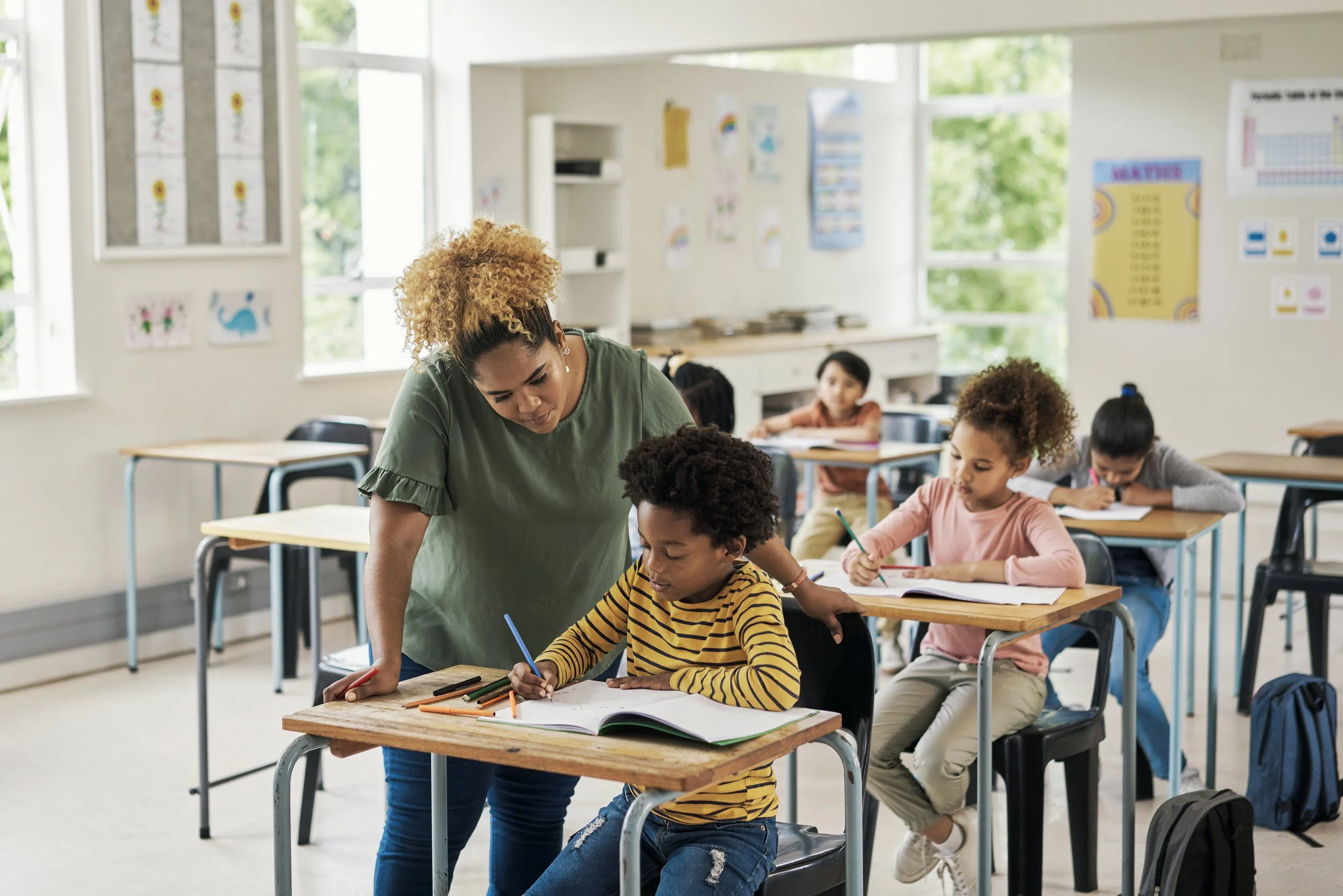 A classroom with young students sitting at desks, writing or drawing, with a teacher assisting a student. The room has large windows, educational posters, and artwork on the walls.