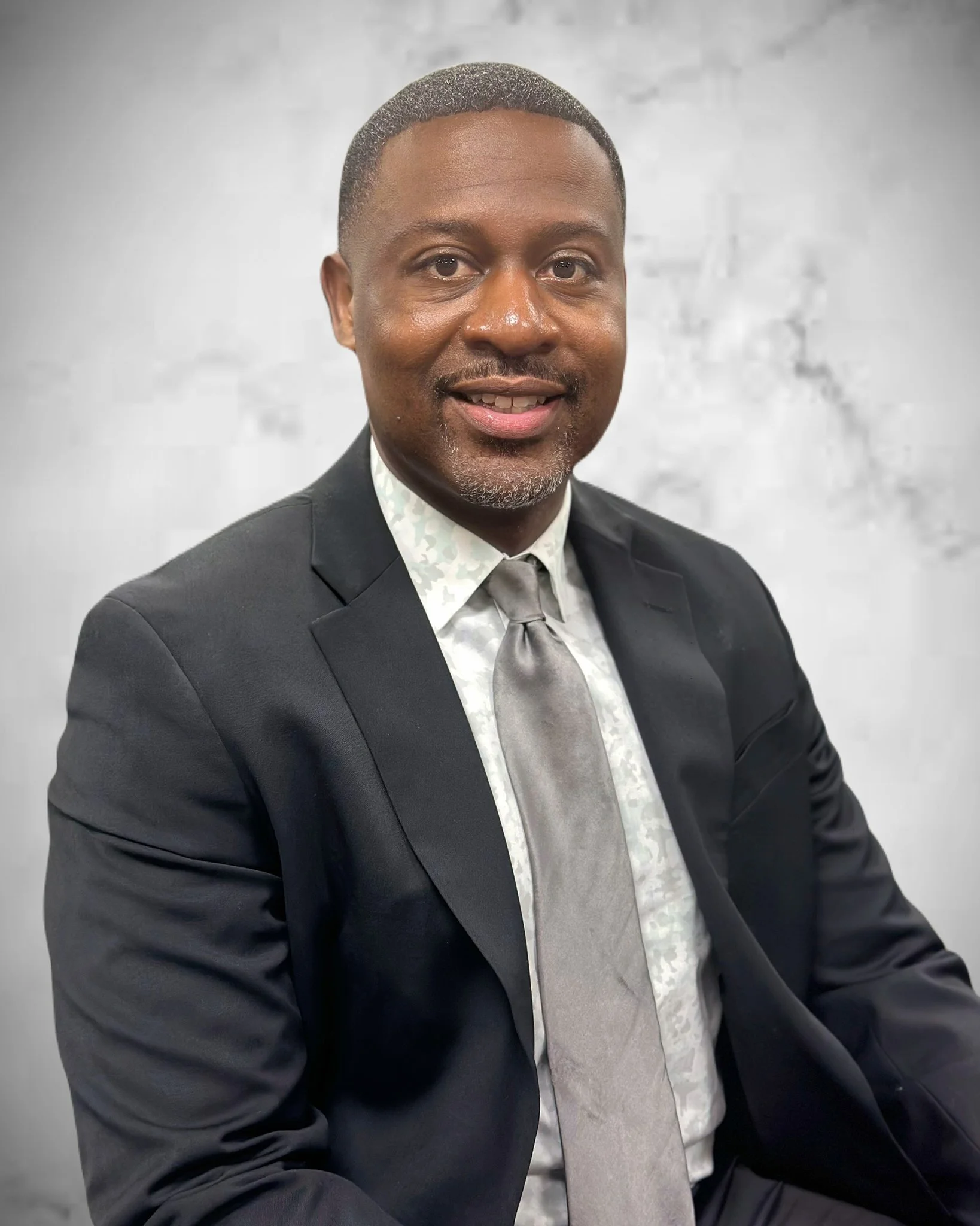 A man in a black suit and patterned shirt with a tie, smiling, against a light-colored brick wall background.