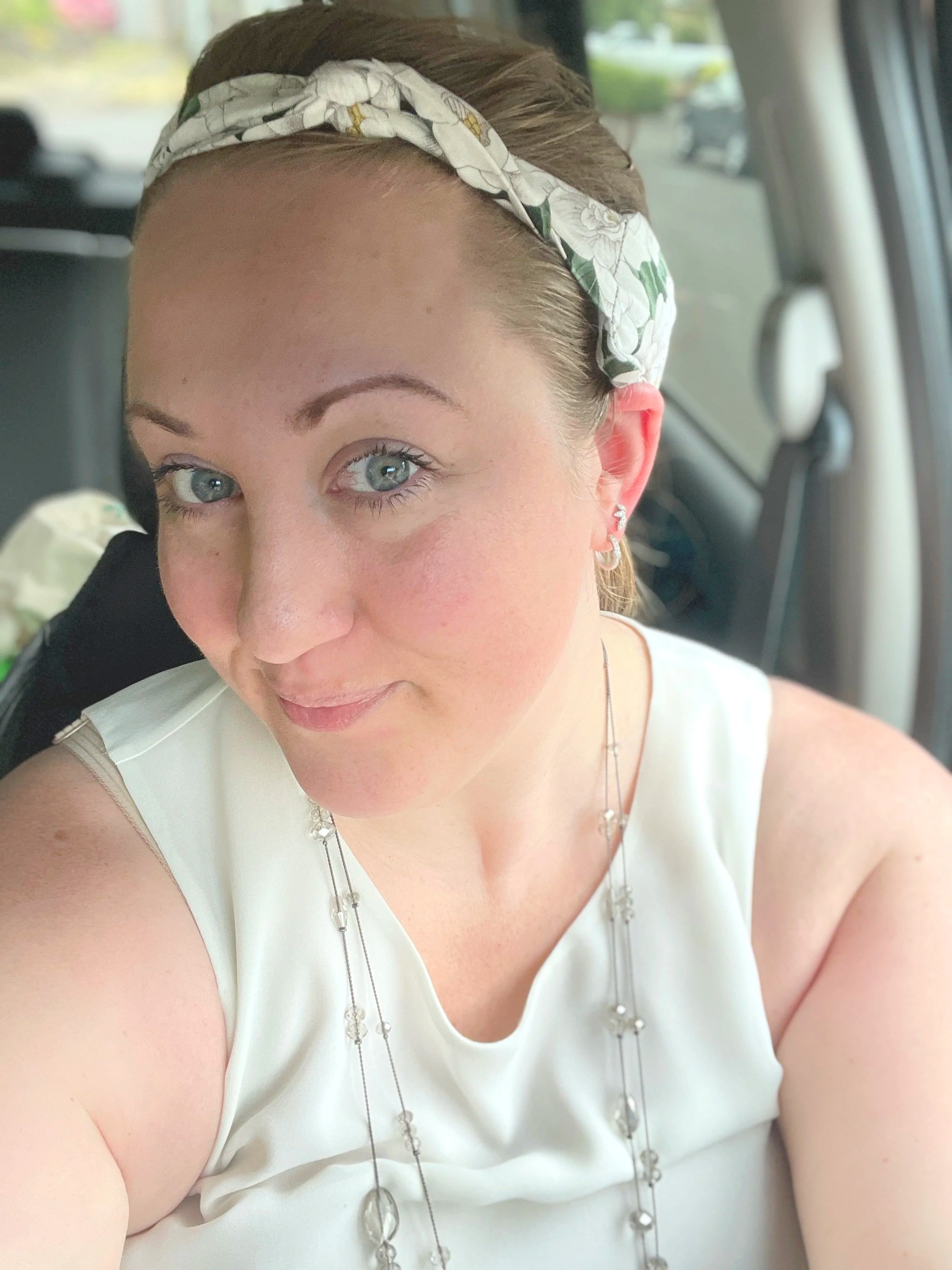 Close-up of a woman with blue eyes, wearing a white Necksicle with a floral pattern, earrings, and a white sleeveless top, sitting inside a vehicle.