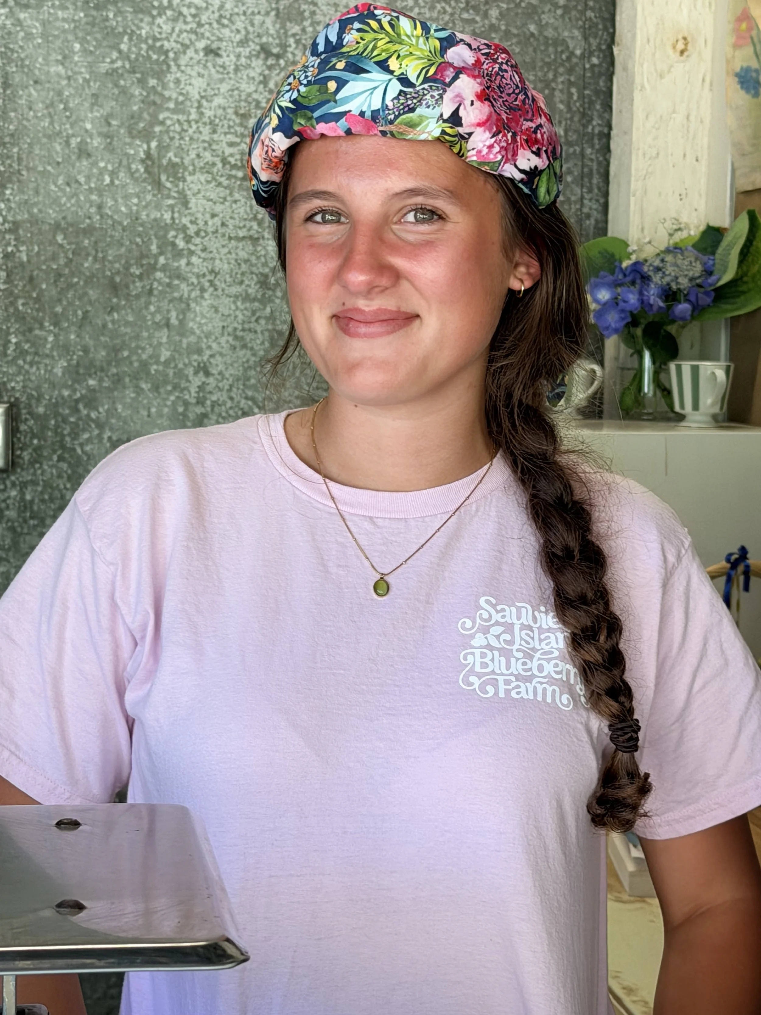 A woman with a long braided hair wearing a colorful tropical print Necksicle and a light pink T-shirt with farm branding, taking a mirror selfie.