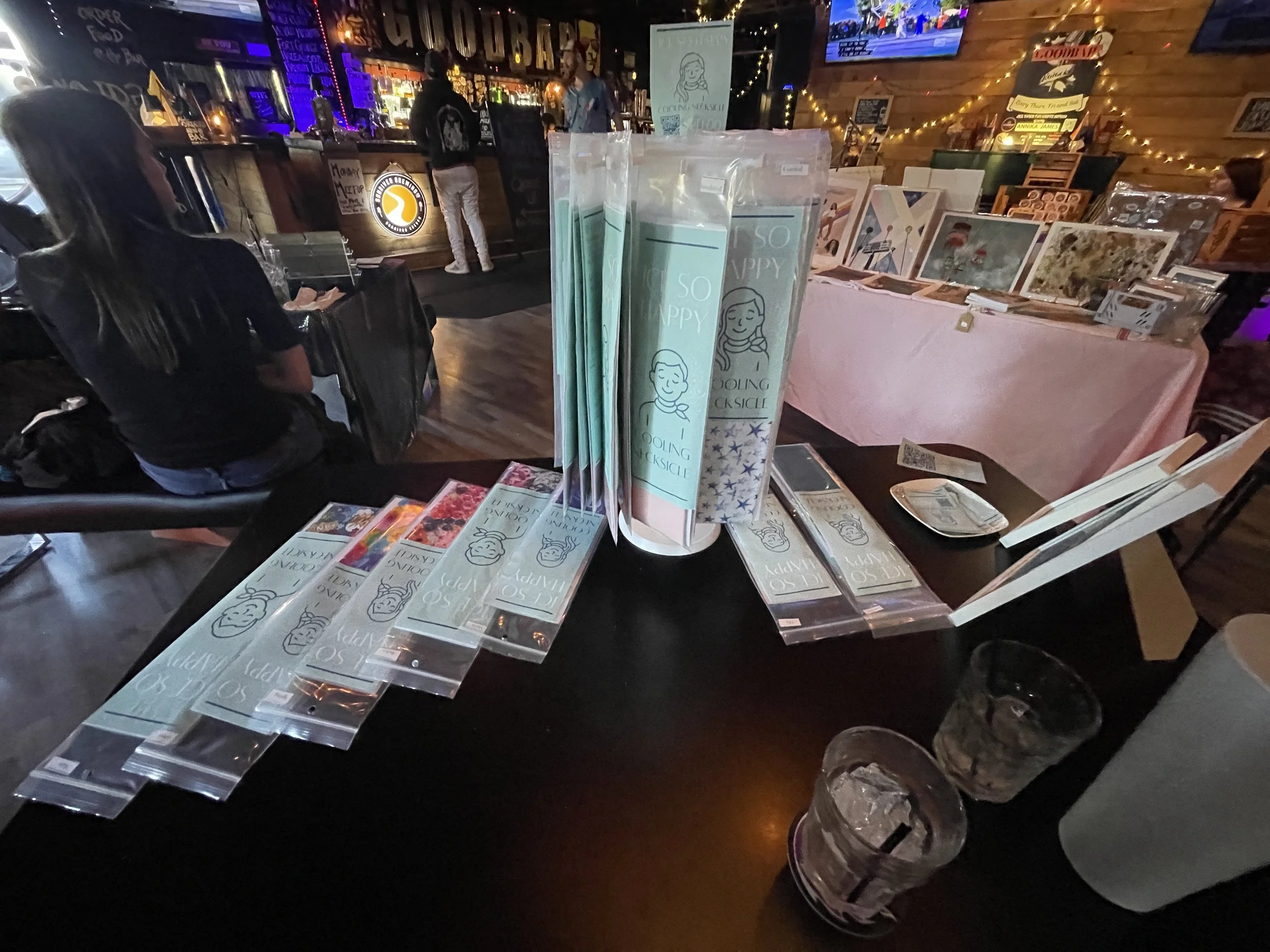 A table at an indoor event displaying various printed materials, including flyers and brochures, with a stack of rolled-up posters or banners featuring a cartoon girl and the words 'SO HAPPY' visible. In the background, there is a bar with people, sh