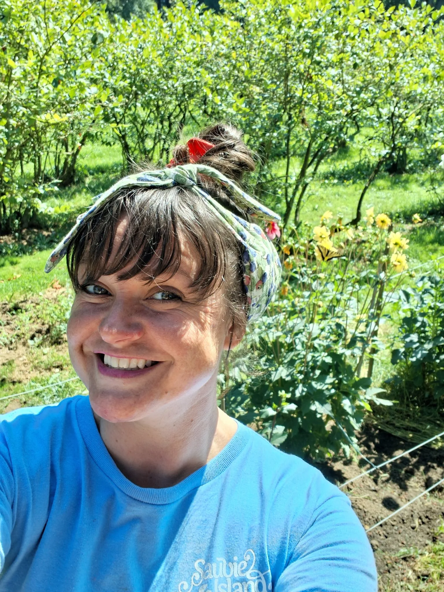 Close-up of a smiling woman wearing a blue shirt and a colorful Necksicle with greenery and yellow flowers in the background.