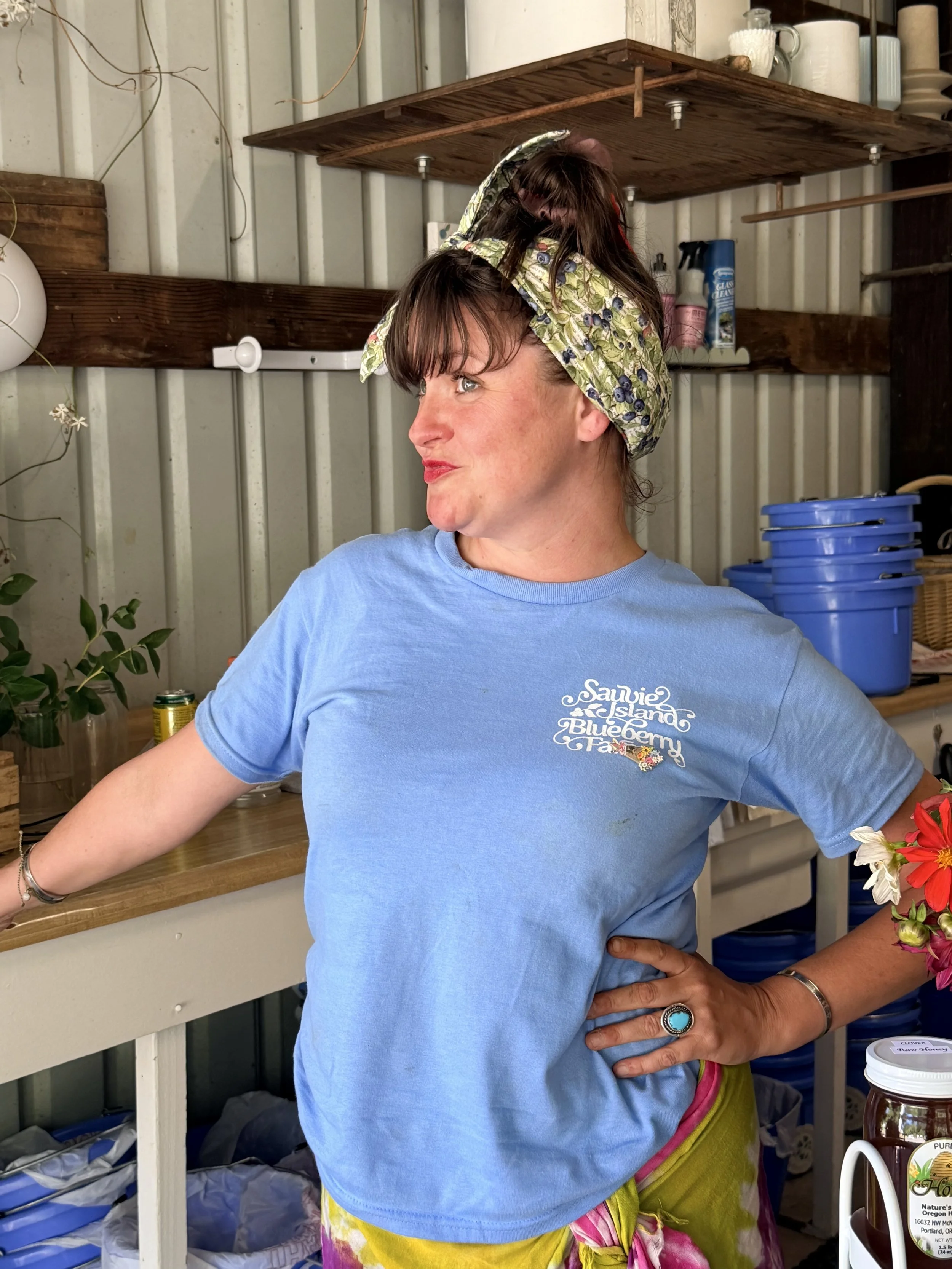 A woman wearing a blue T-shirt with a printed design and bright multicolored skirt, sporting a large floral Necksicle, standing outdoors near a wooden shelf with various household items, including a jar of honey, plants, and containers.