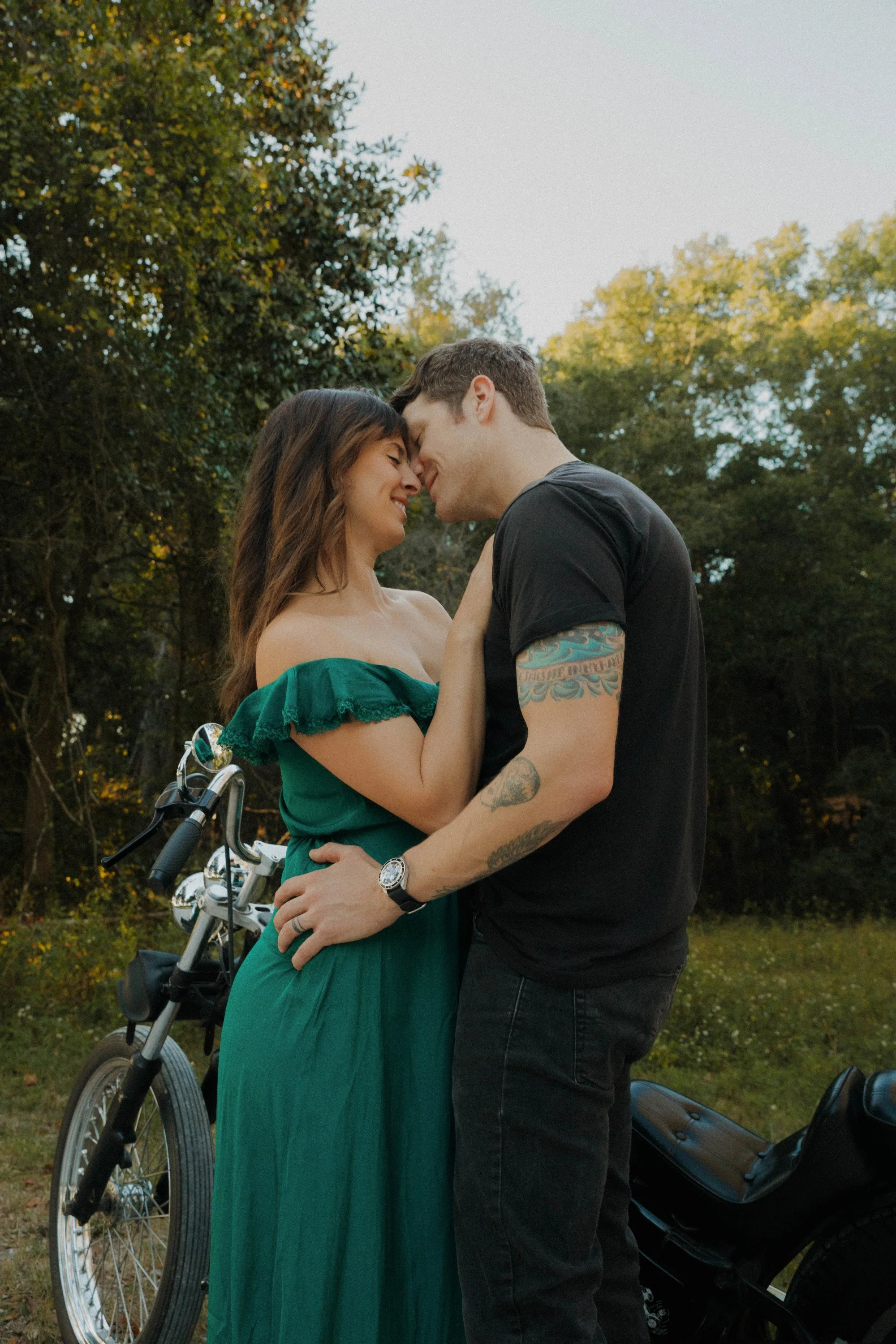 A couple at sunset, touching foreheads and smiling, outdoors with trees in the background, a bicycle with a basket nearby, and a black motorcycle beside them.
