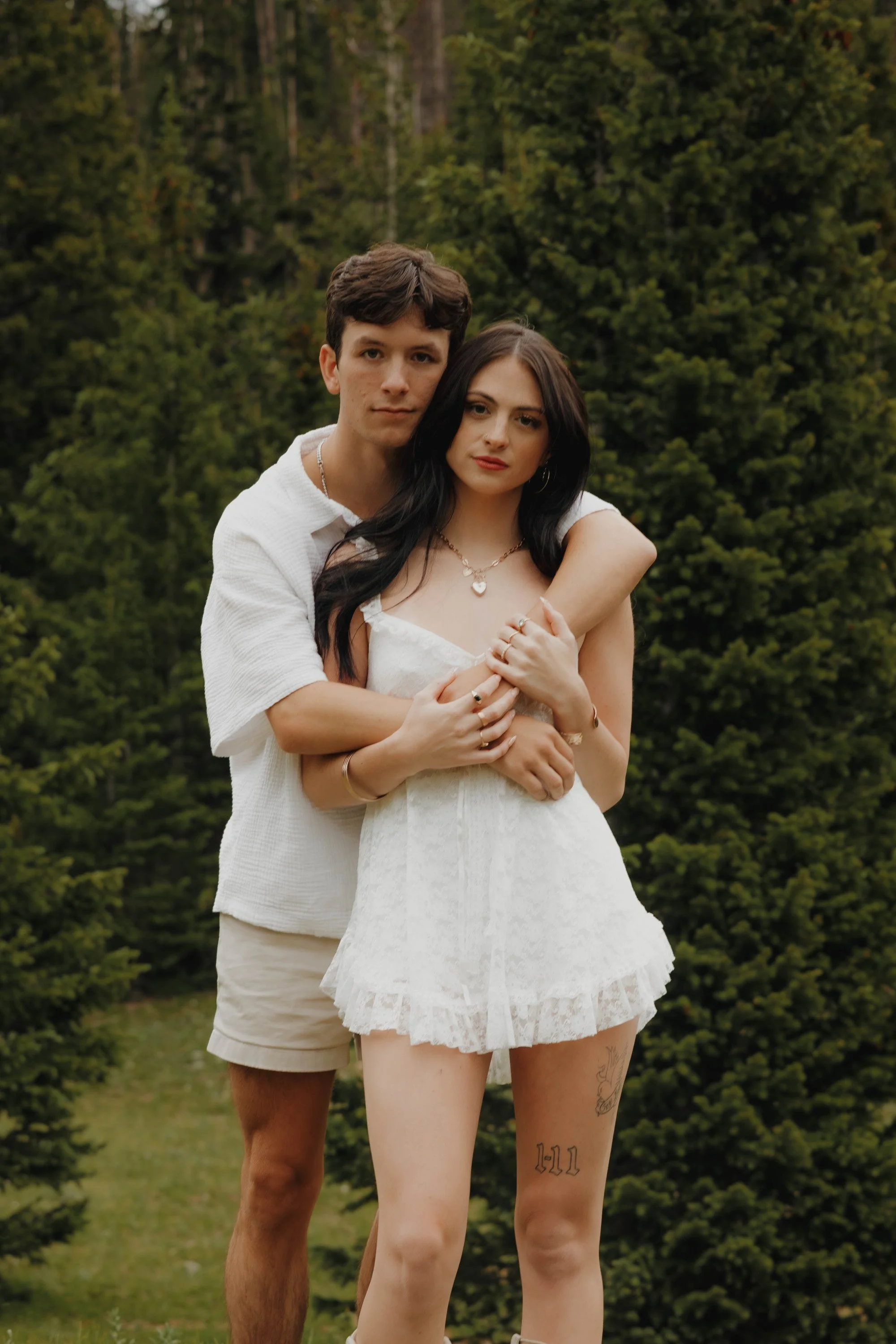 A young couple standing outdoors in front of green trees, with the woman in a white dress and the man in a white shirt and beige shorts, embracing each other.