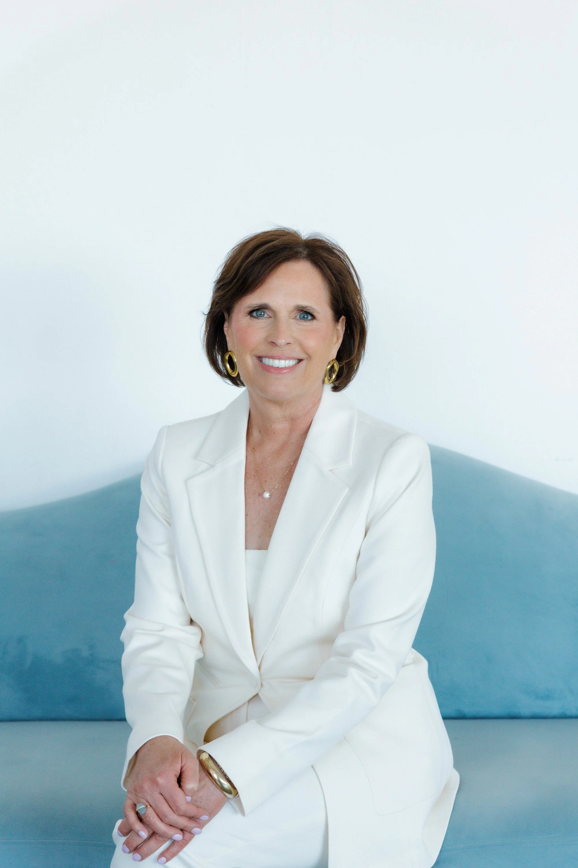 A woman with short brown hair wearing a white suit and gold jewelry, sitting against a plain light background.