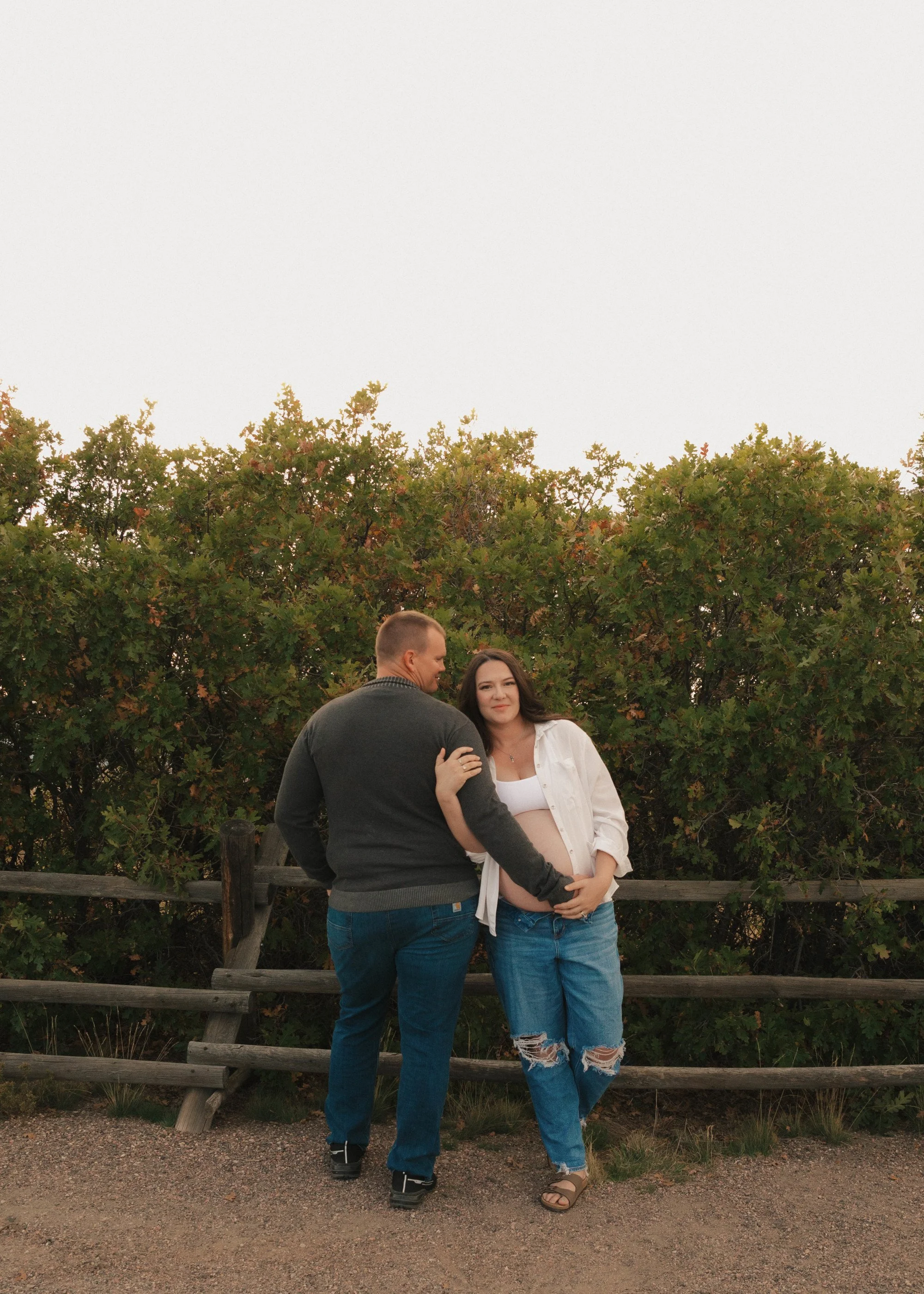 A pregnant woman and a man standing together outdoors in front of trees and a wooden fence, with the woman resting her hand on her belly as they both smile.