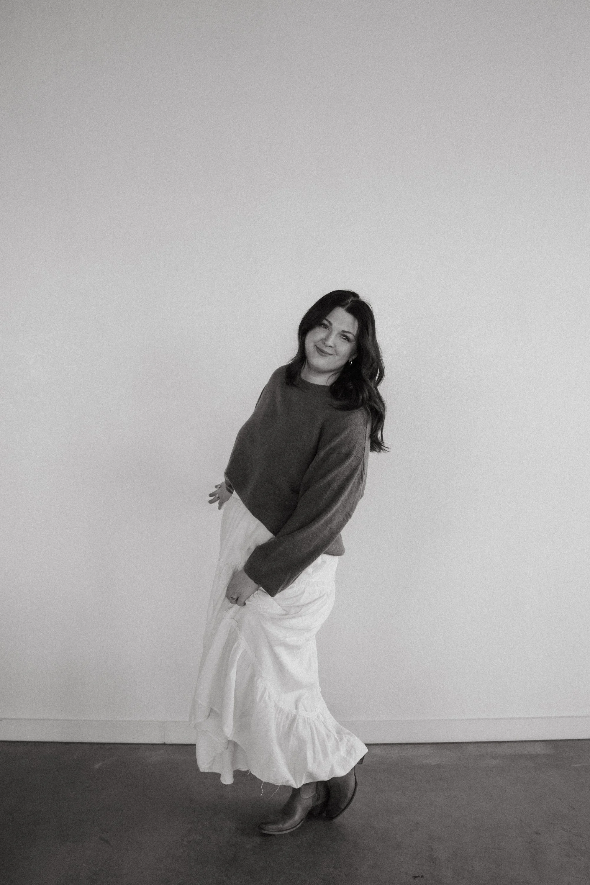 A woman with dark, wavy hair wearing a long-sleeved, loose-fitting shirt and a flowing ankle-length skirt, standing indoors against a plain wall. She is smiling and facing the camera.