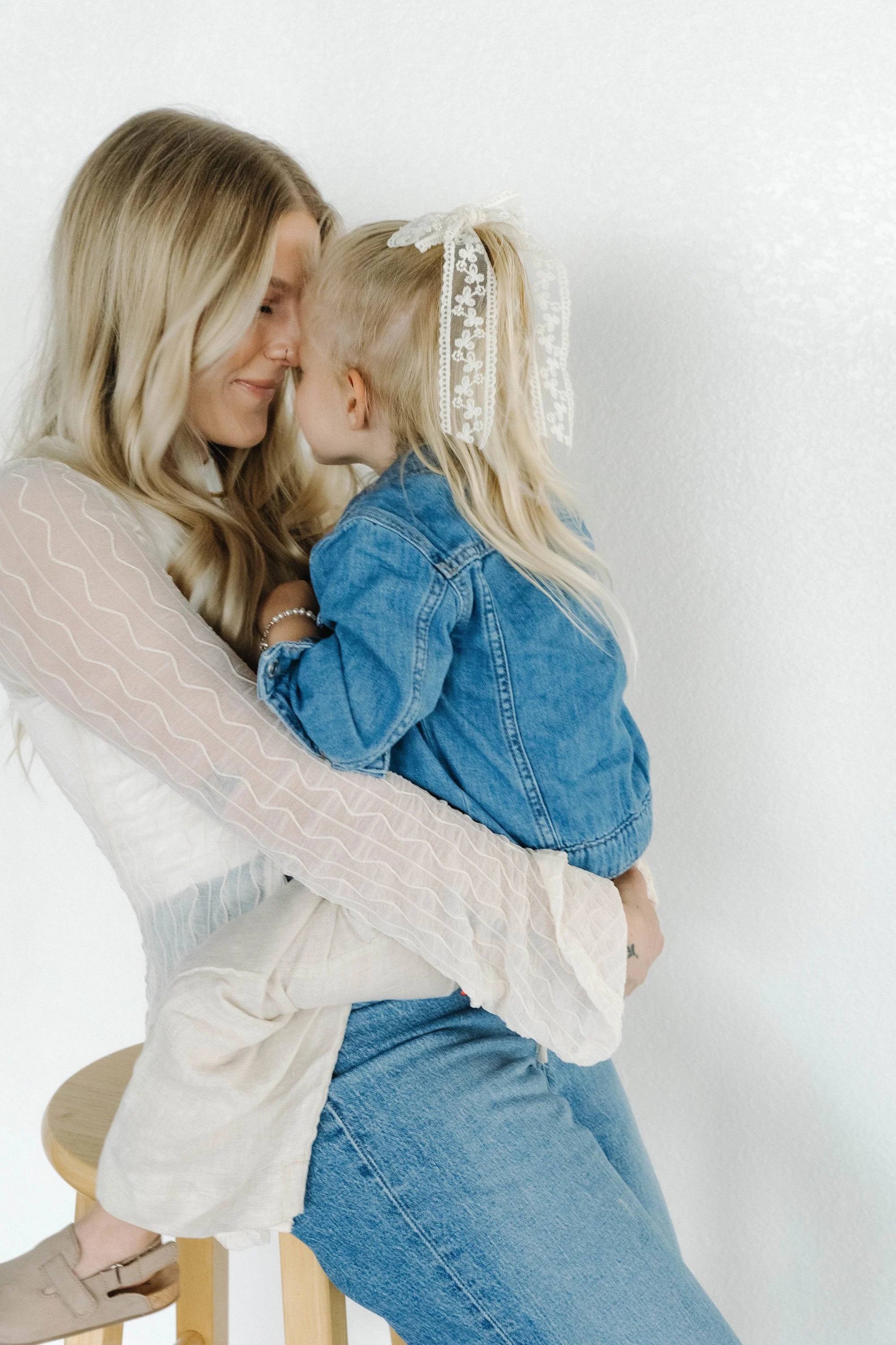 A woman with long blonde hair and a young girl with long blonde hair and a lace bow, sharing a close moment against a plain white wall. The woman is sitting on a stool, wearing a light pink blouse, and holding the girl, who is wearing a denim jacket 