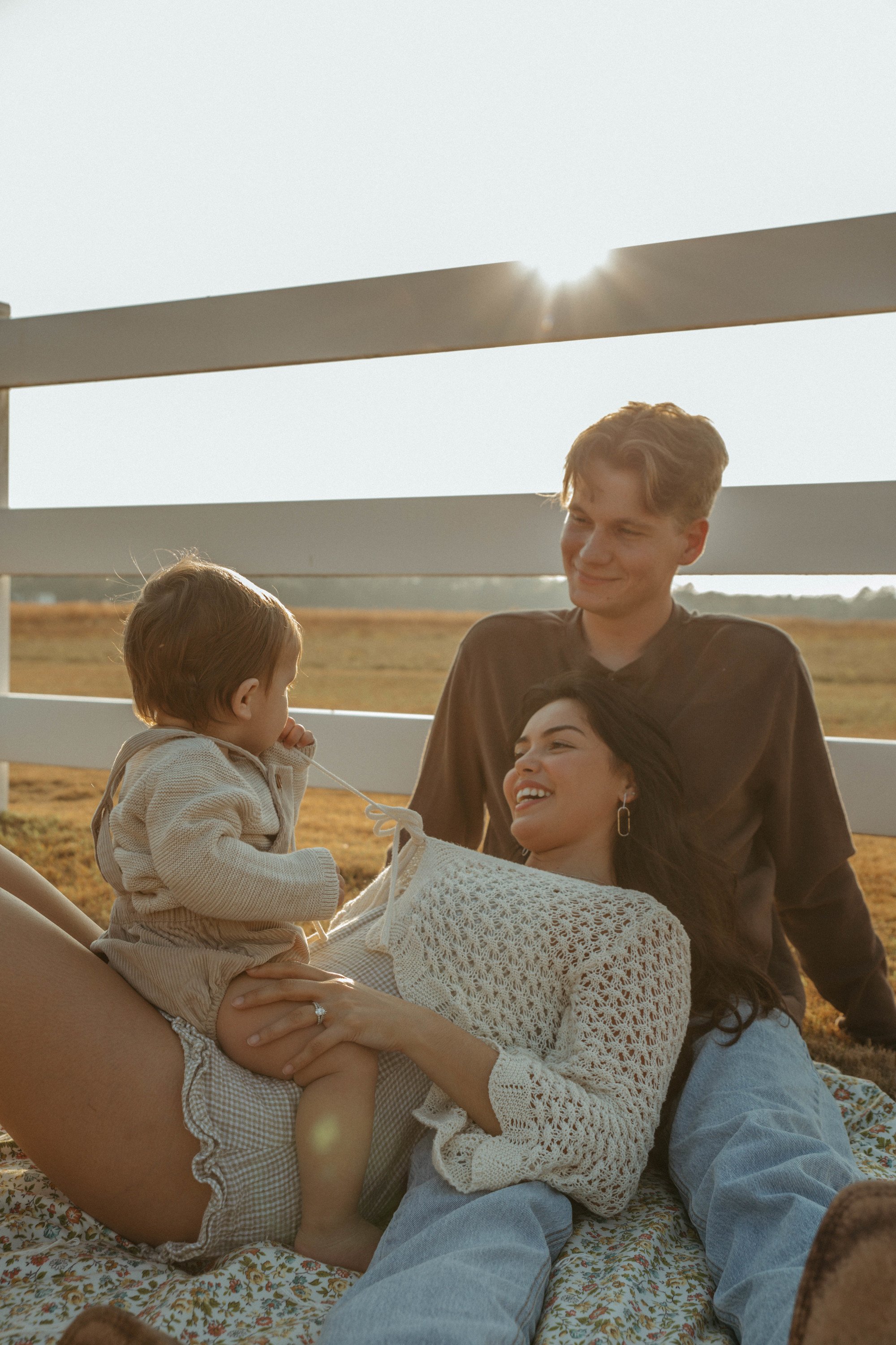 A family of four enjoying a moment together outdoors at sunset, with a white fence and open field in the background.
