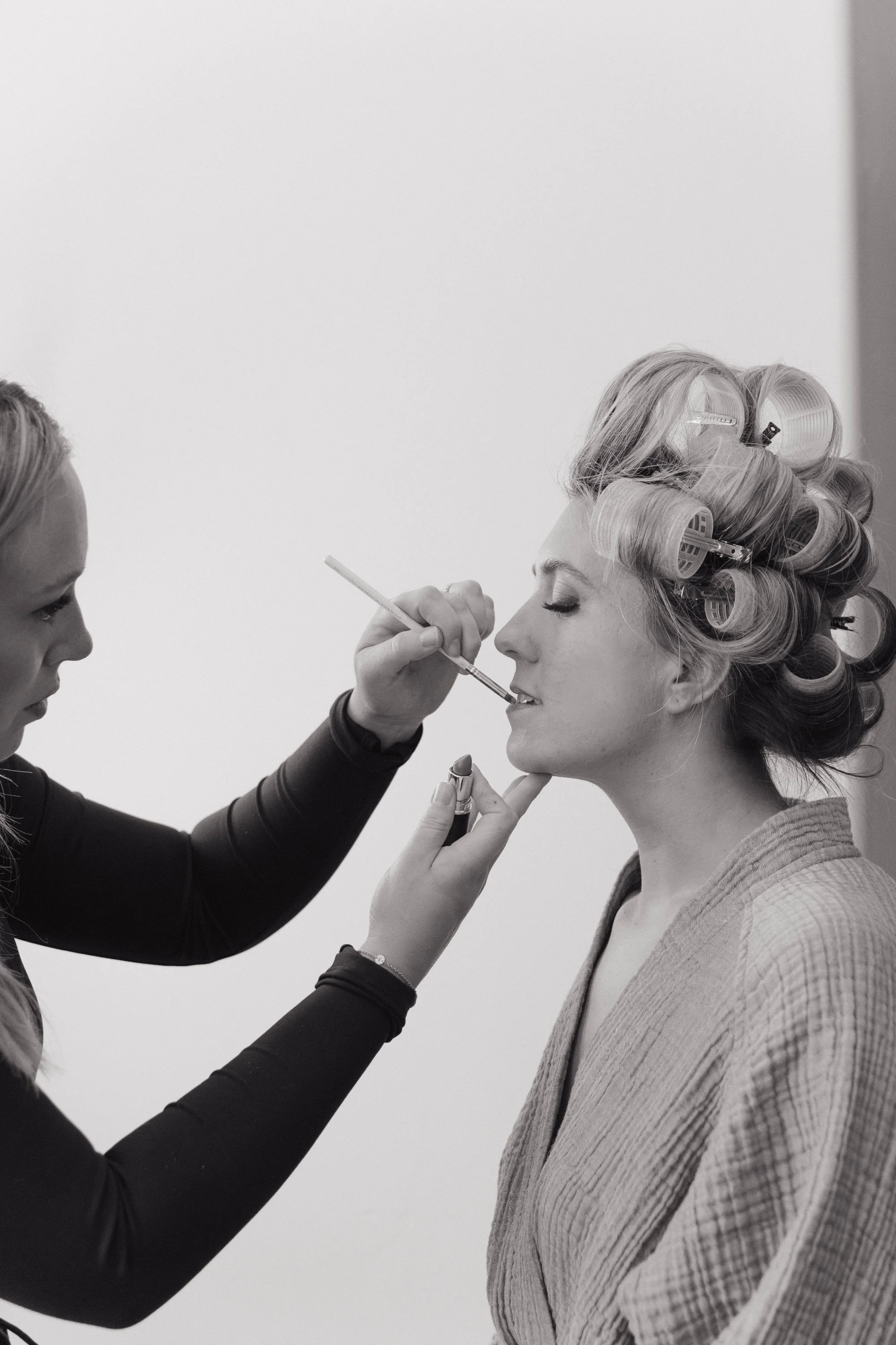 A woman with curlers in her hair having makeup applied by a makeup artist.