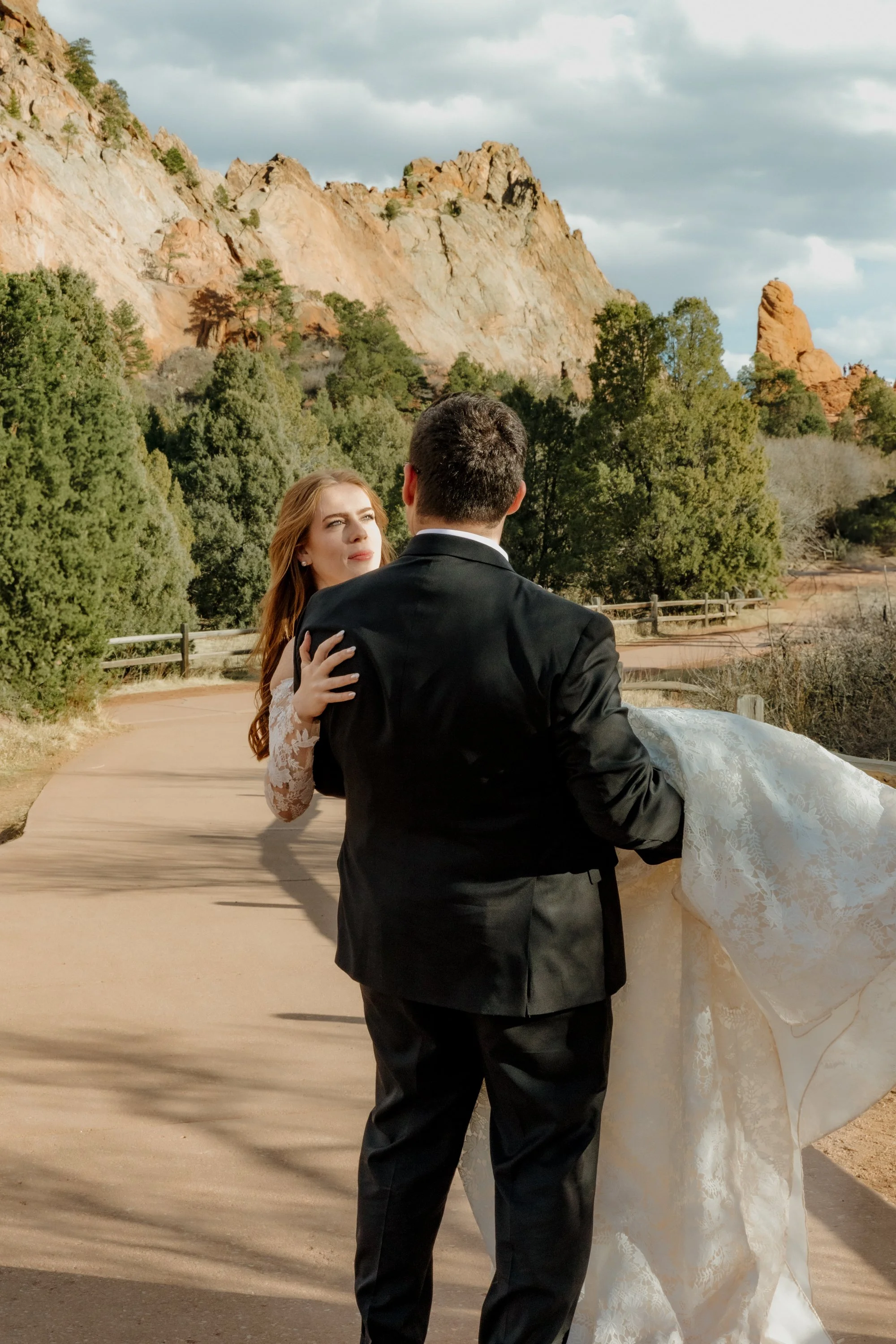 A man in a black suit and a woman in a wedding dress dancing outdoors with a mountainous landscape in the background, including trees, rocky formations, and a cloudy sky.