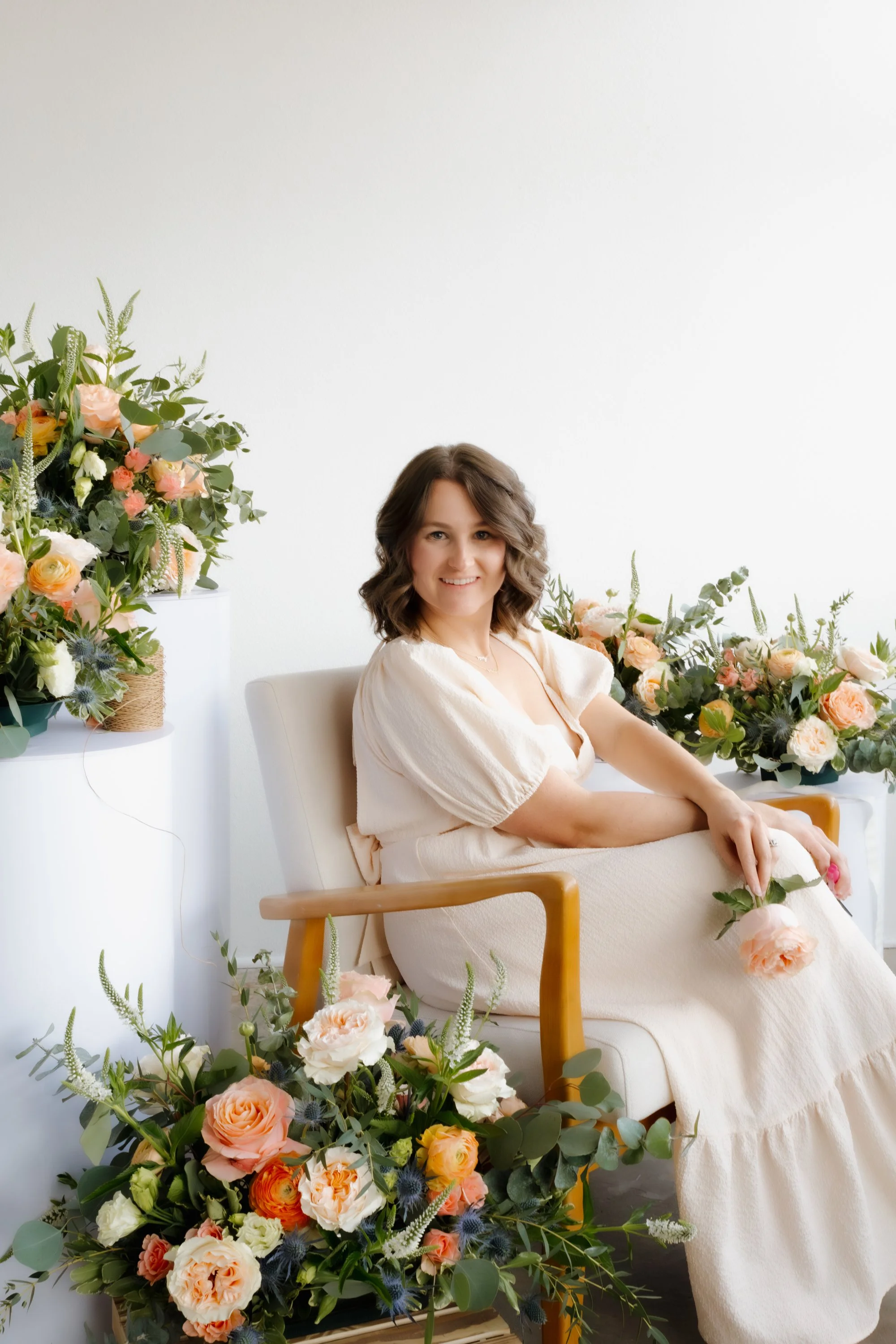A young woman with shoulder-length brown hair smiling while sitting on a beige armchair surrounded by large floral arrangements with peach, white, and yellow roses, and green foliage in a bright, white room.