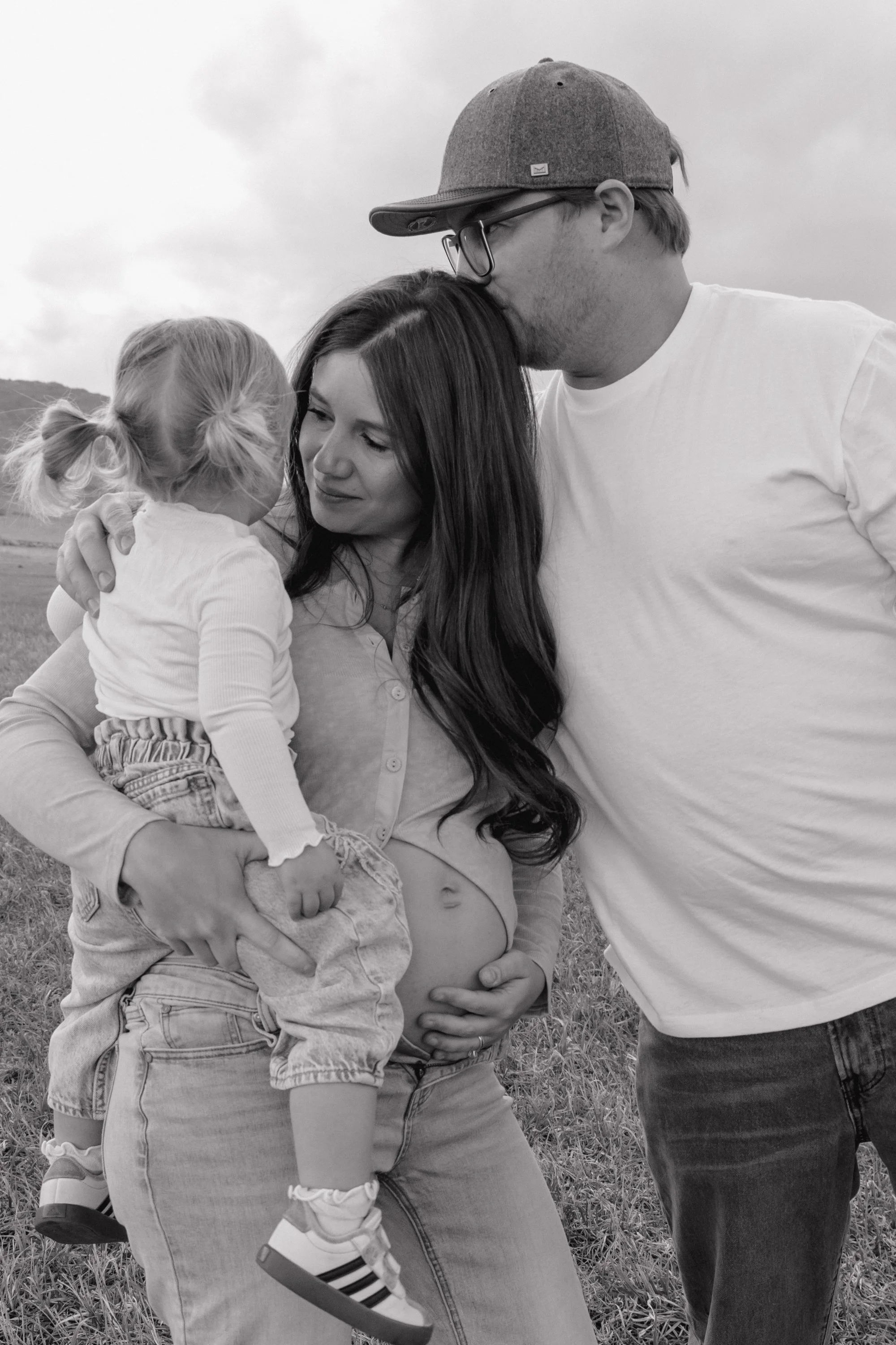 A family outdoors, with a pregnant woman holding a young girl in her arms, a man kissing the woman's forehead, all standing on grass, in a natural setting with cloudy sky.