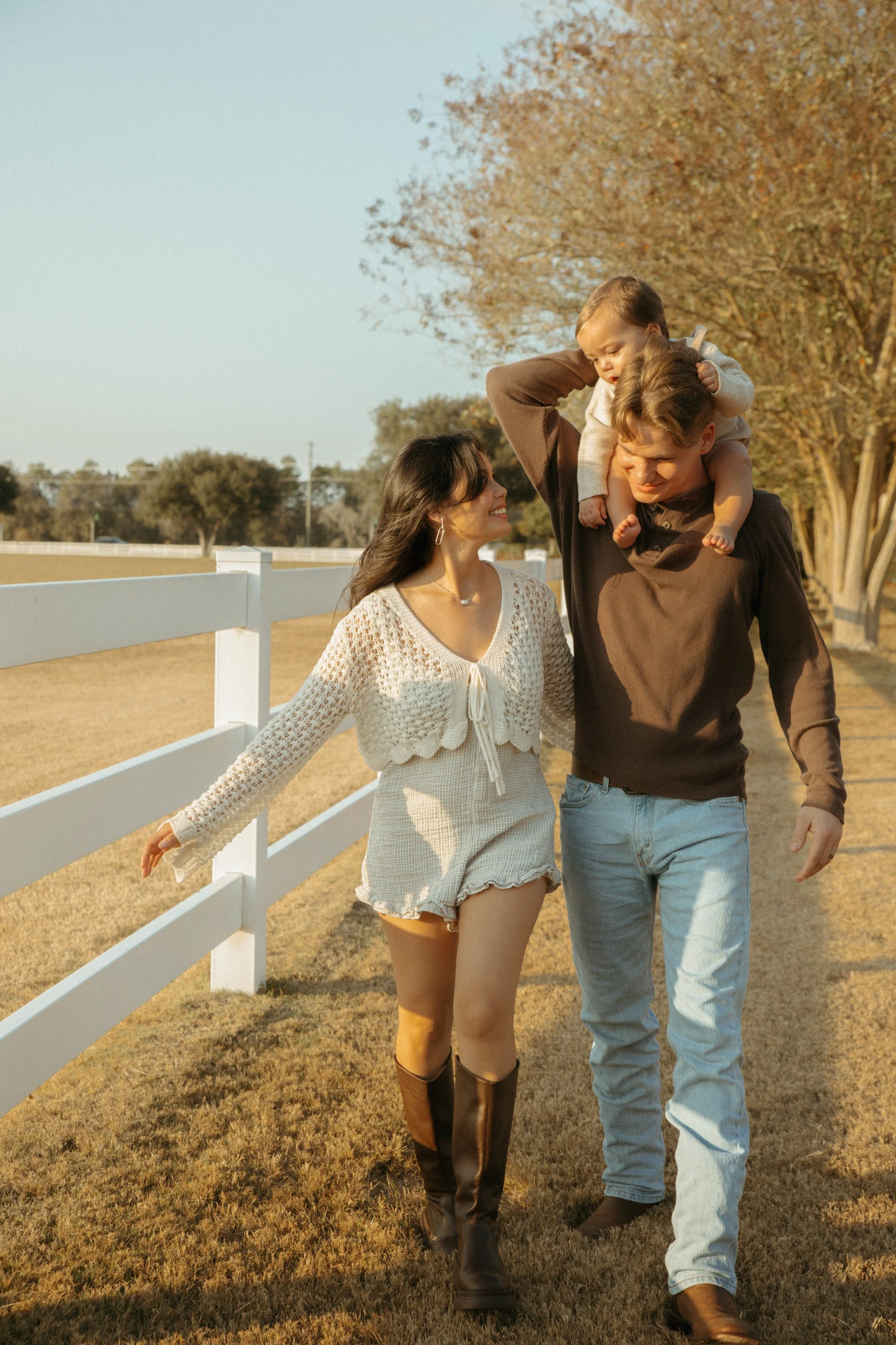 A family of three walking outdoors near a white fence, with trees in the background and sunlight casting a warm glow.