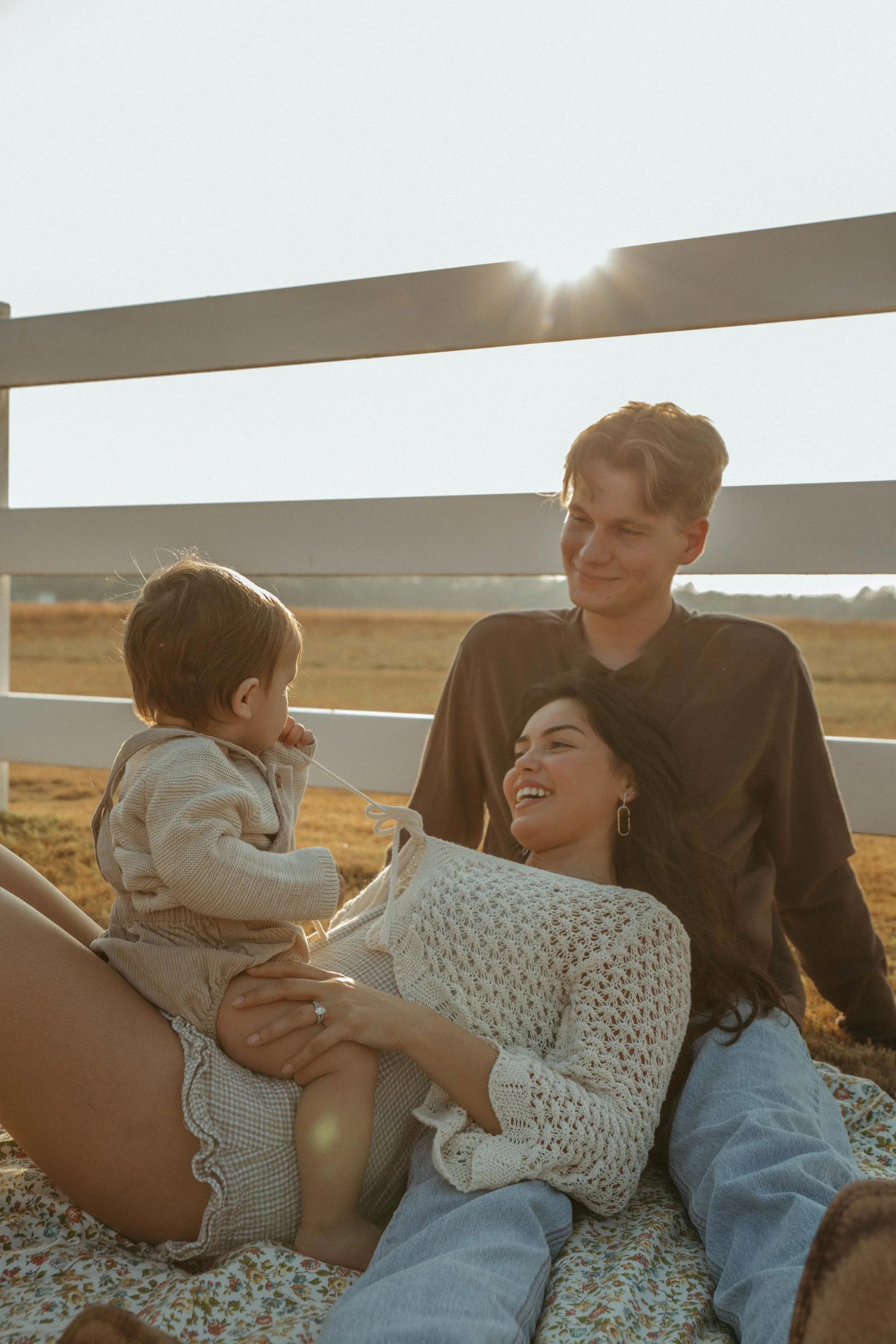 A woman and a man sitting outdoors with two children, one girl and one boy, enjoying a moment together during sunset on a farm or open field, with a white fence in the background.