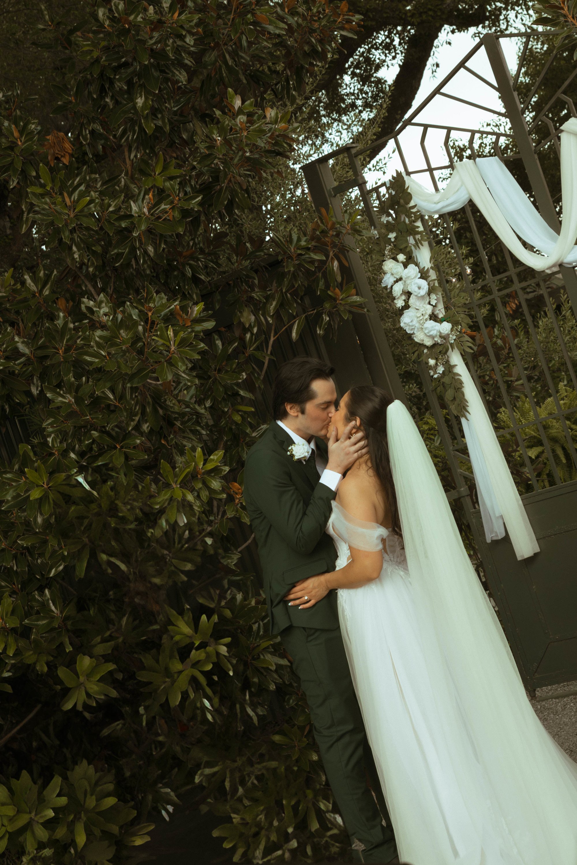 A bride and groom kissing outside in front of a floral wedding arch at nighttime, with leafy bushes and a tree in the background.