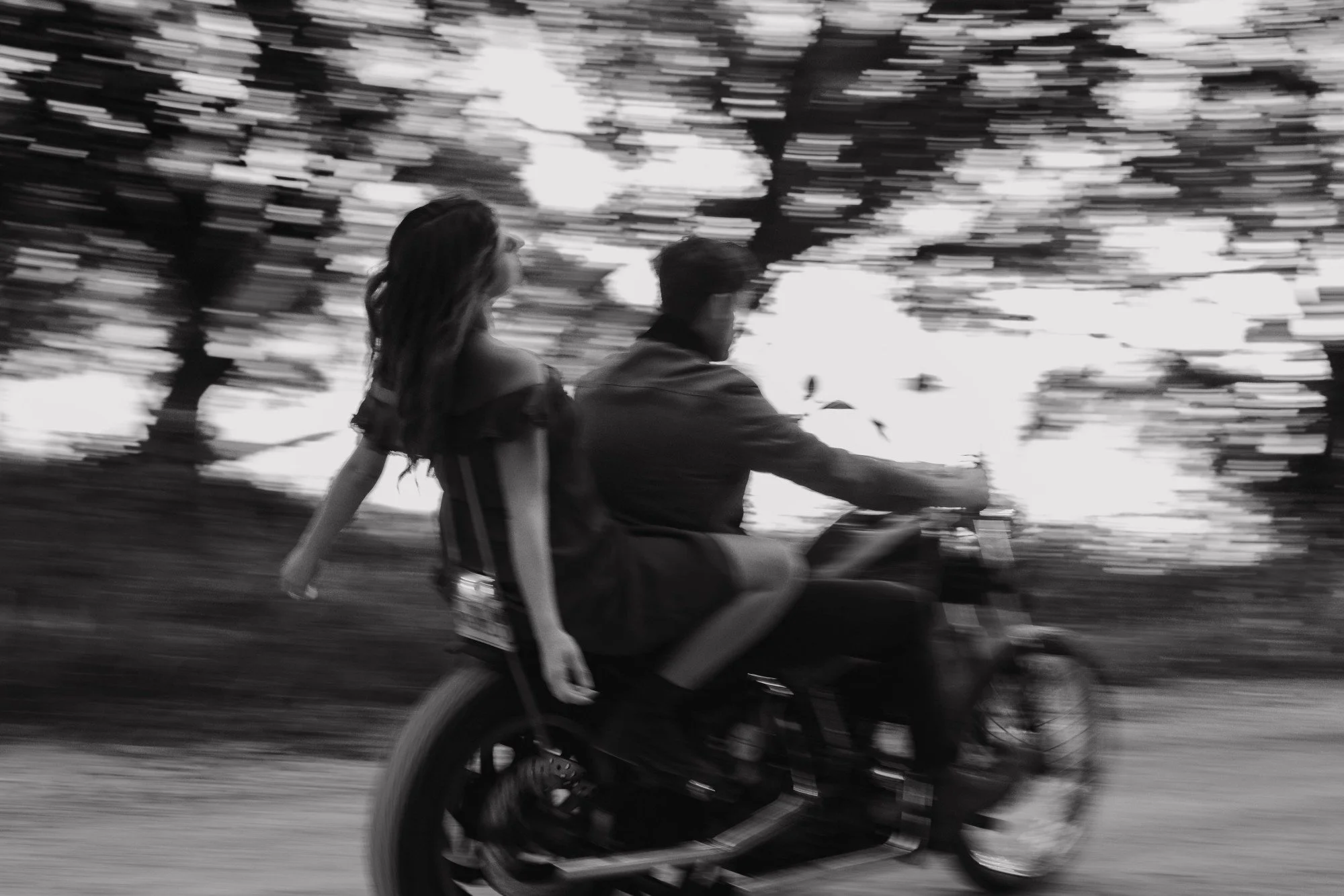 Black and white photo of a man and woman riding a motorcycle outdoors. The woman has long hair and is wearing a dress, while the man is wearing a jacket. The background shows blurred trees and sky, indicating motion.