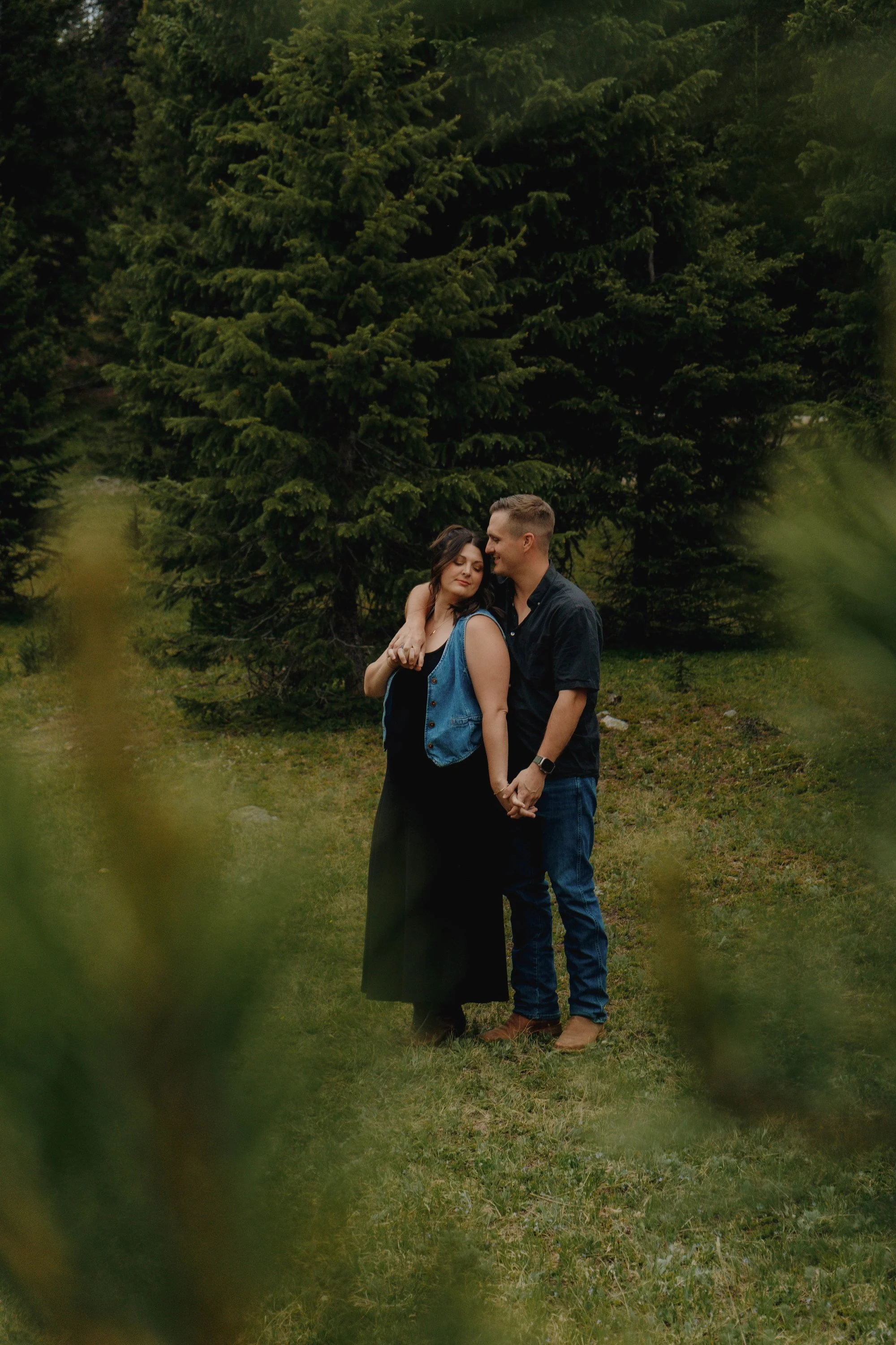 A young couple stands close together outdoors, holding hands and smiling, surrounded by tall green trees.