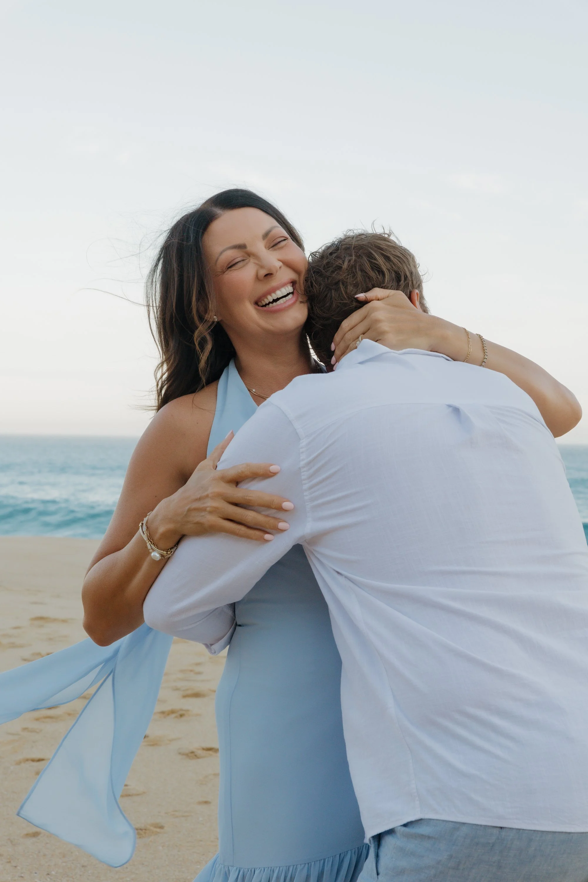 A woman with dark wavy hair in a blue dress is hugging a man wearing a white shirt on the beach. They are smiling, and the ocean and sky are visible in the background.
