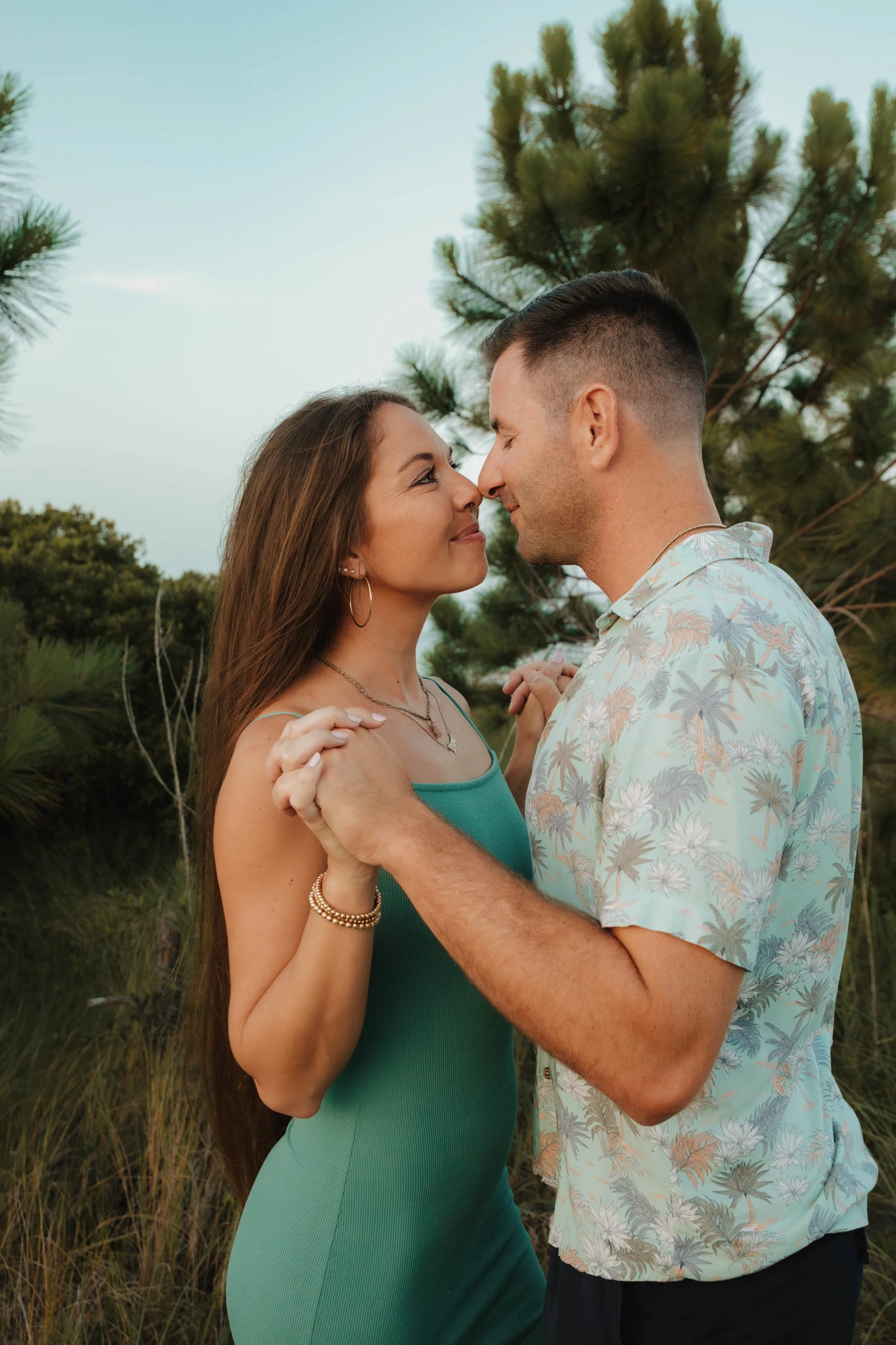 A couple dancing closely outdoors, smiling at each other, with trees and blue sky in the background.